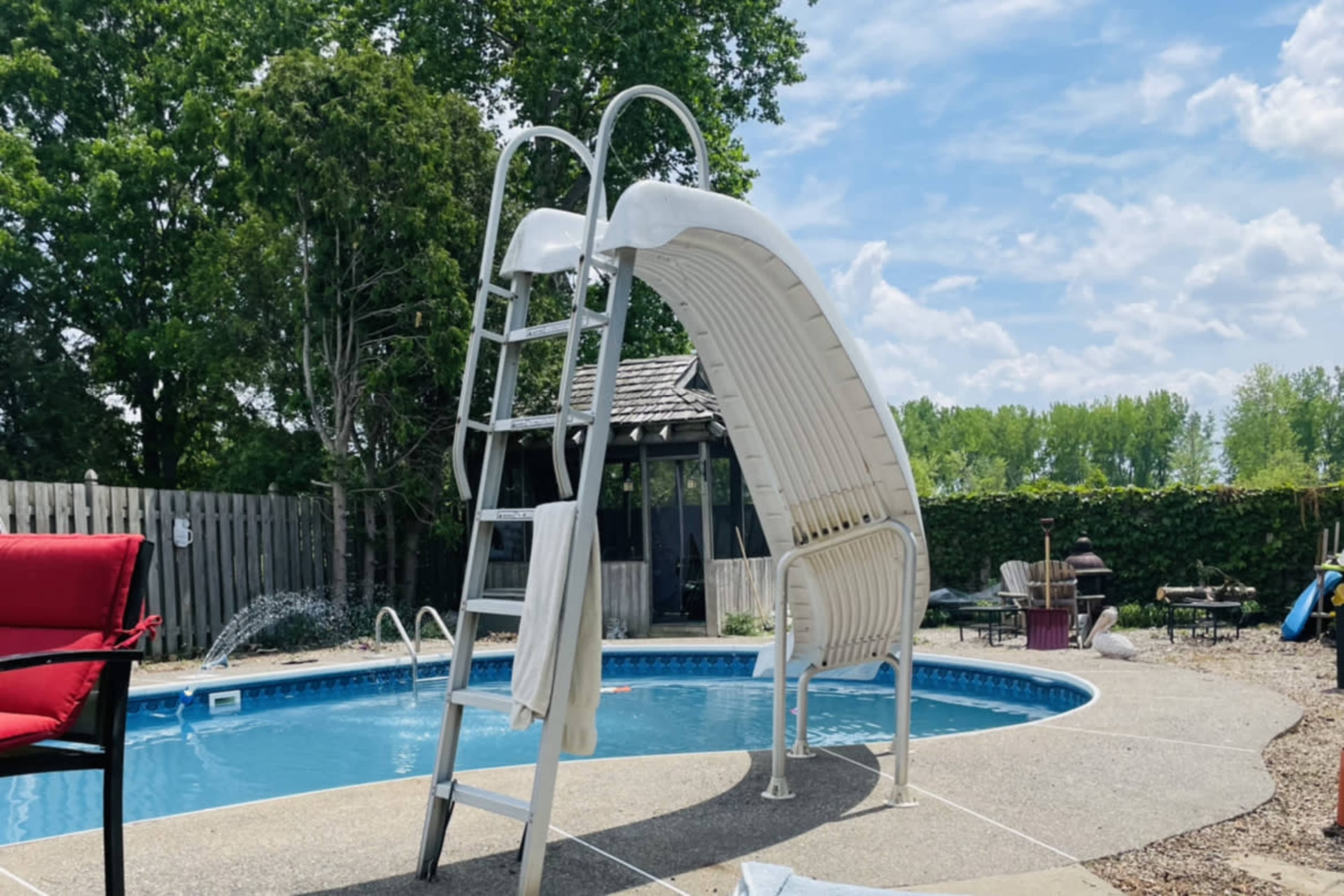 A white water slide stands beside a circular swimming pool in a backyard, with trees and seating areas in the background.