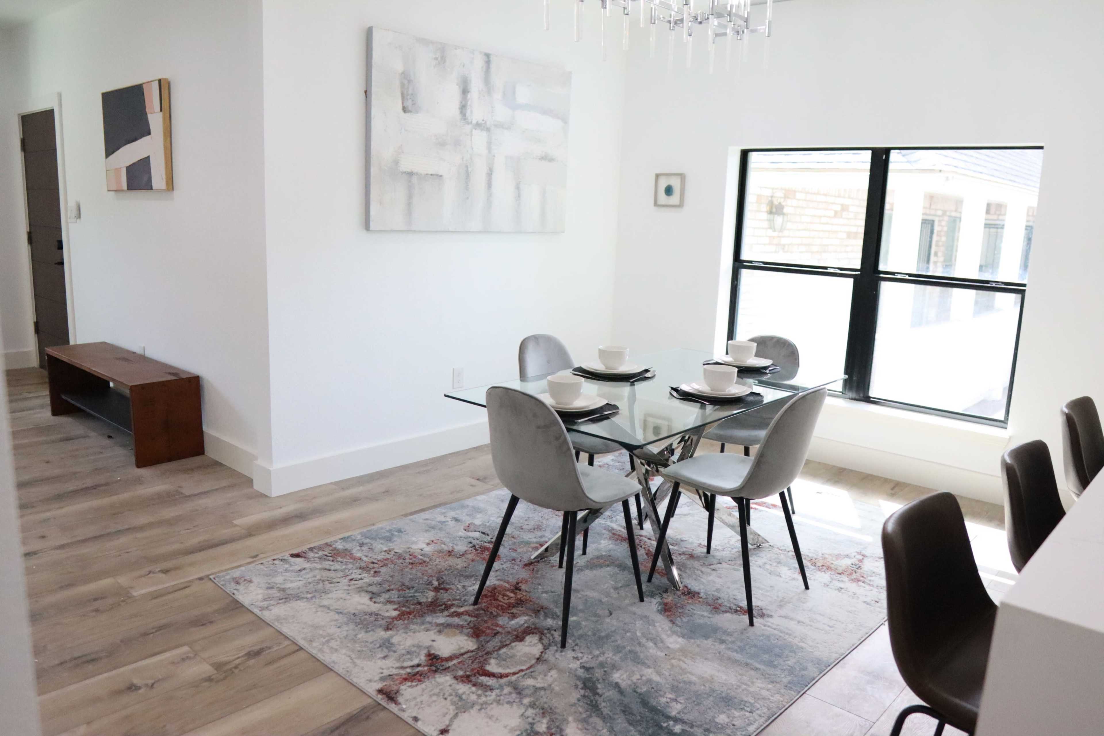 A modern dining area features a glass table set for six, surrounded by gray chairs, and is illuminated by natural light from a large window.