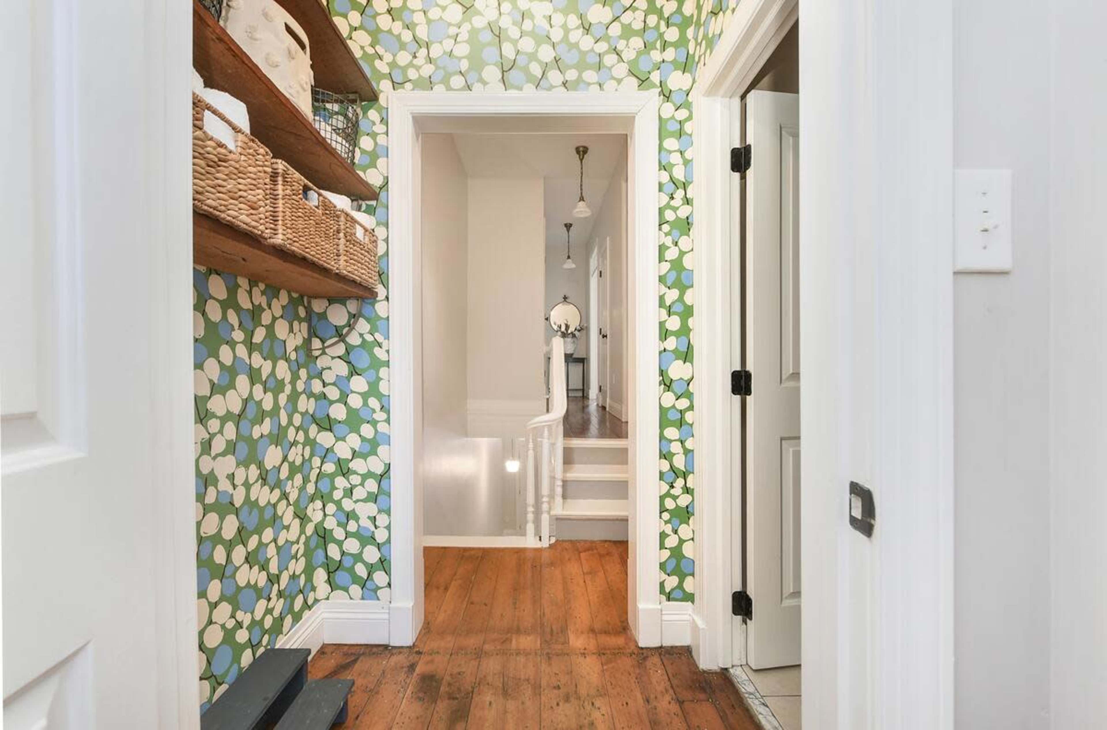 The image shows a hallway with patterned wallpaper, wooden flooring, and doors leading to a staircase and other rooms.