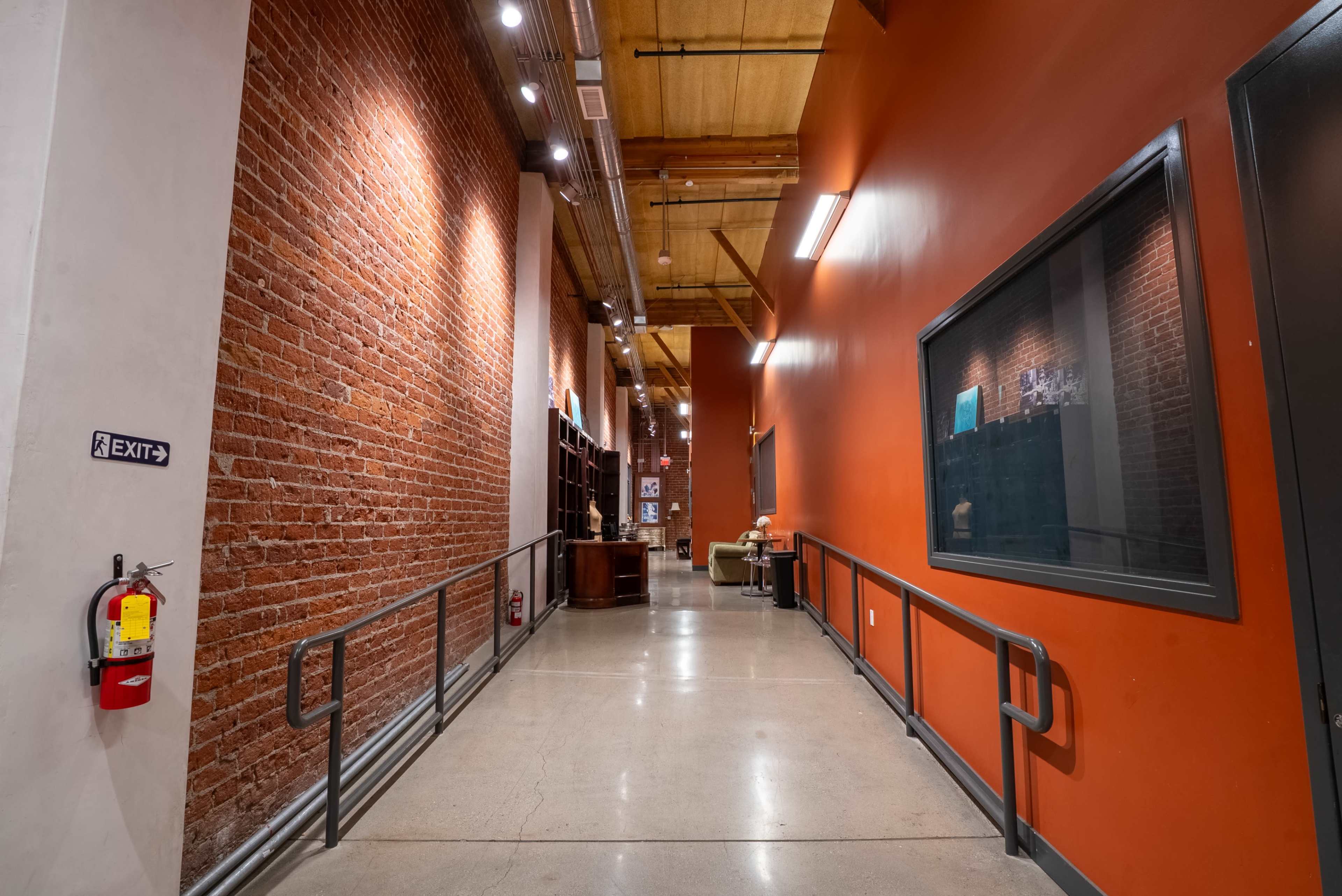 The image shows a narrow corridor with red walls and exposed brick, leading to a workspace with furniture at the far end.