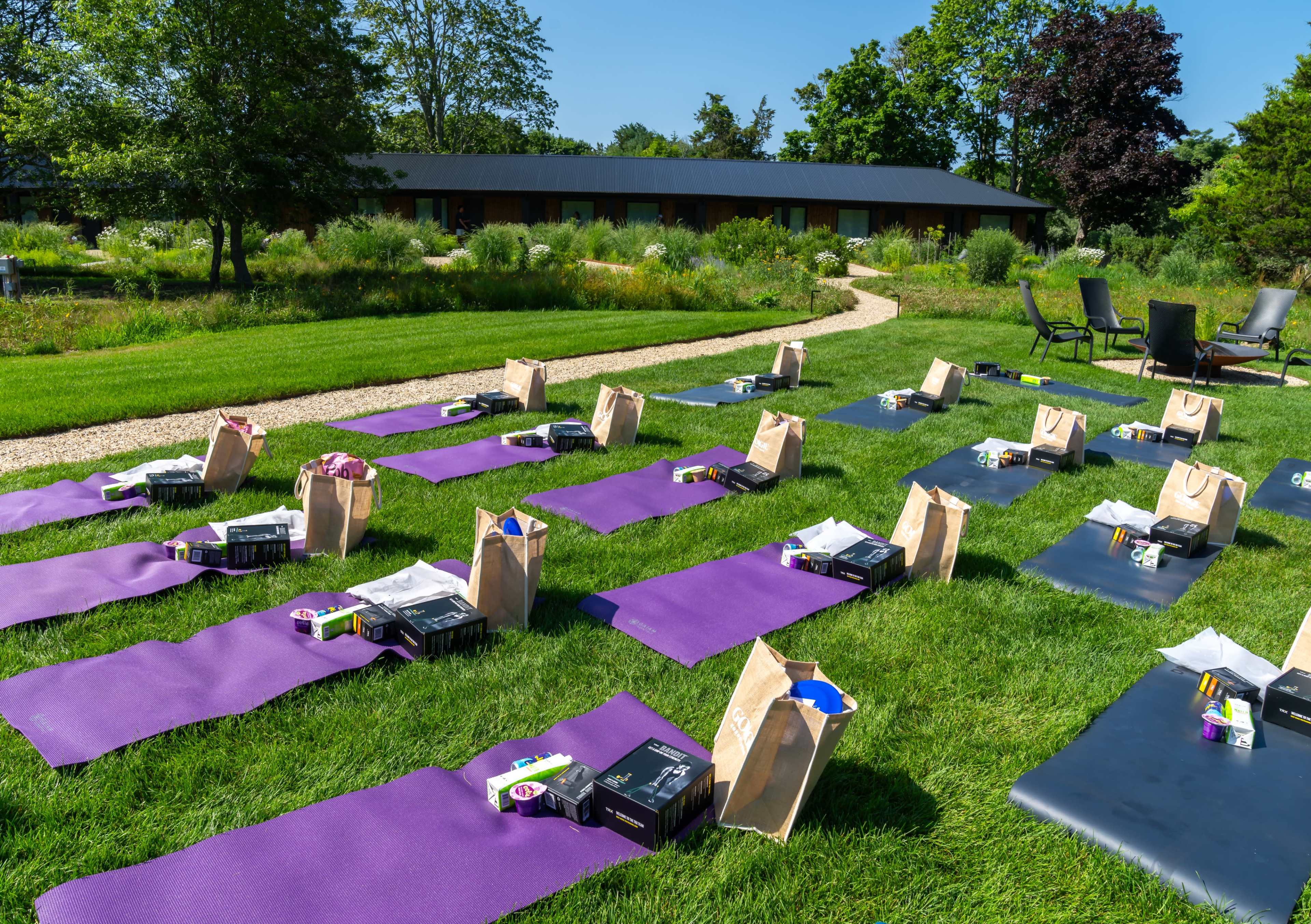 The image shows a row of yoga mats arranged on a grassy area, each accompanied by a bag, with a pathway and trees in the background.