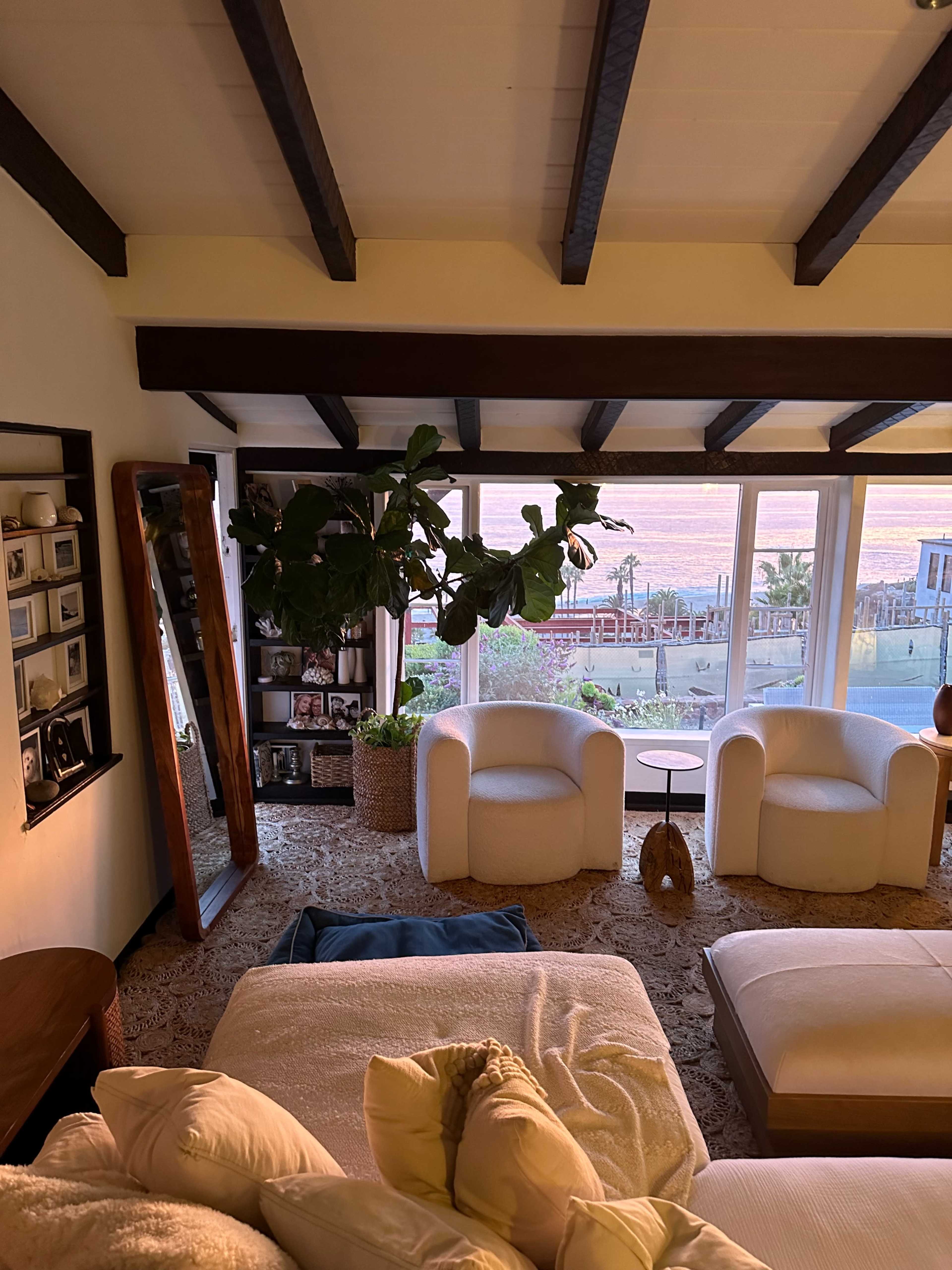 A cozy living room features two armchairs, a small side table, a large mirror, and a tall plant, with large windows showcasing a view of the outdoors.