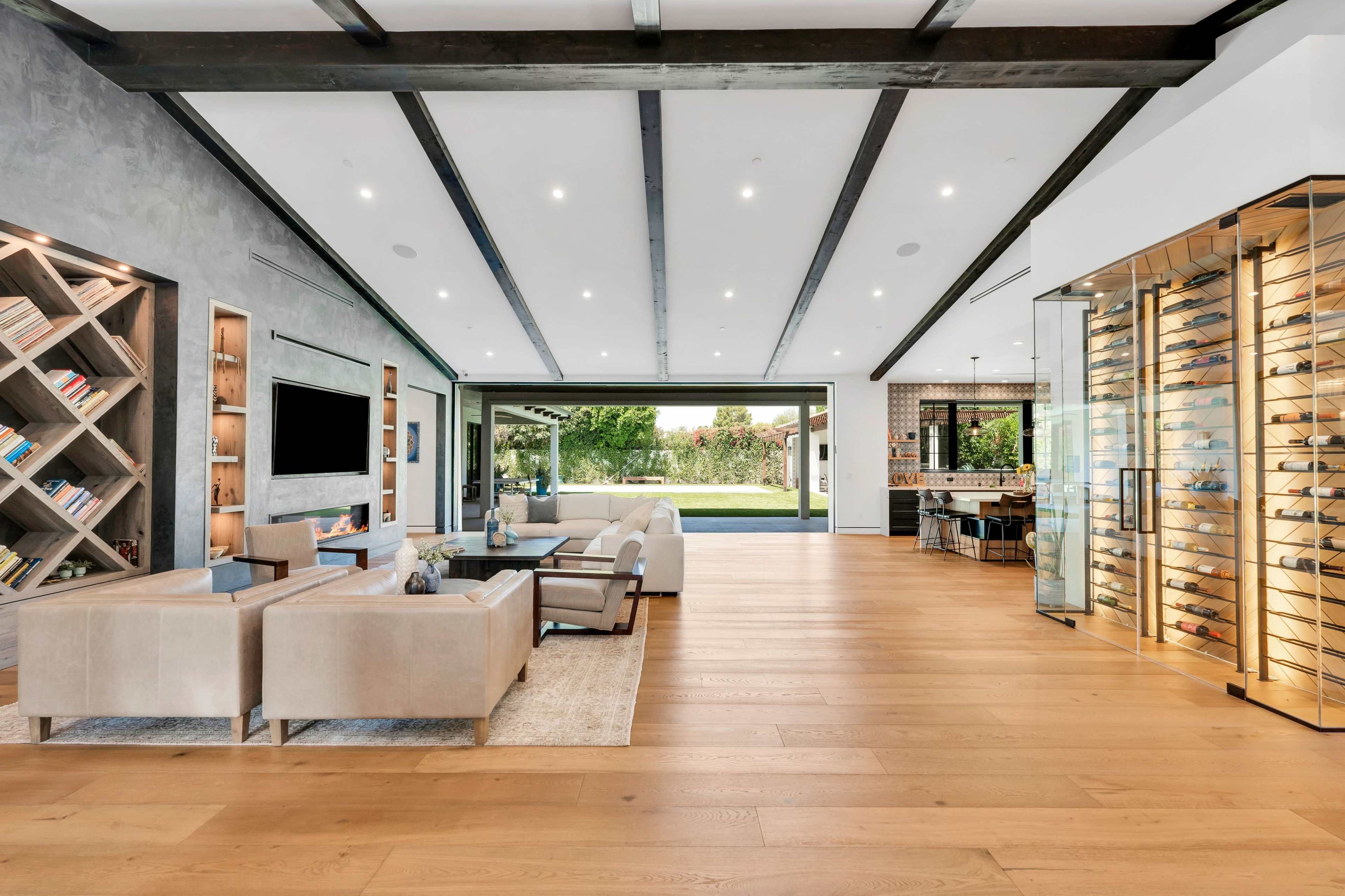 A spacious living area features a sectional sofa, a television mounted on a gray wall, and a glass wine display alongside an open kitchen area that leads to an outdoor view.