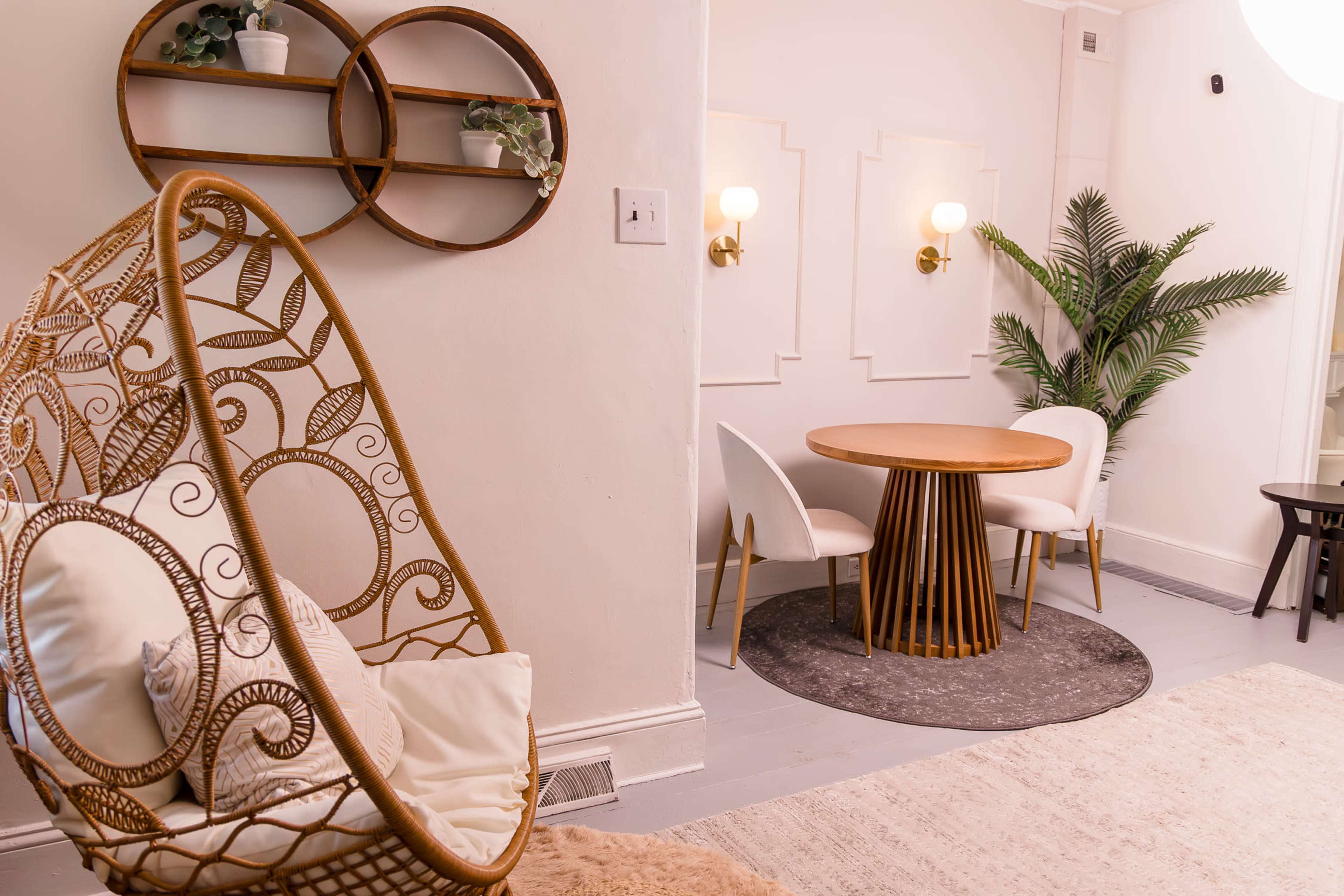 The image shows a cozy interior featuring a rattan hanging chair, a round wooden dining table with white chairs, and a decorative plant beside the table.