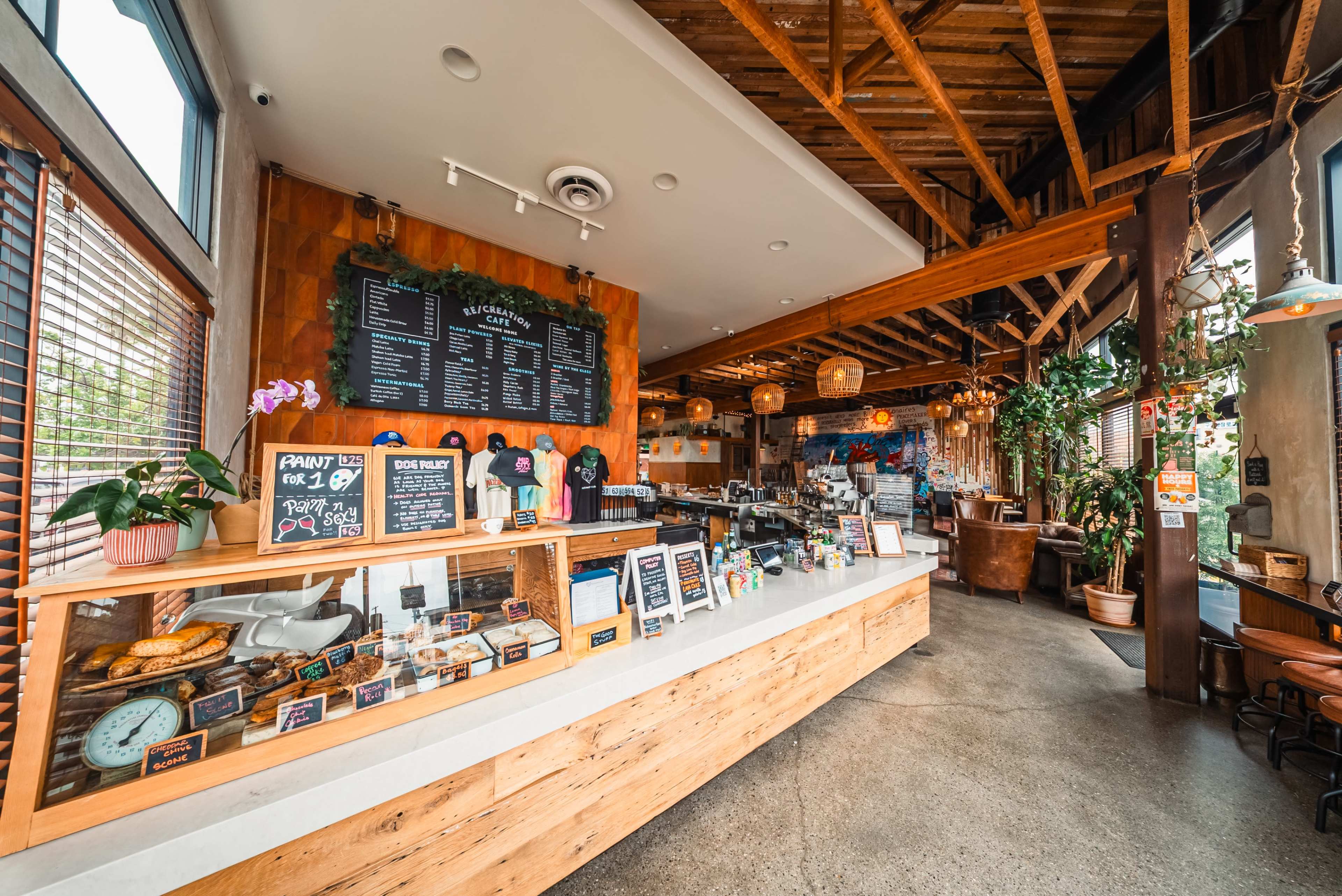 The image shows a coffee shop interior featuring a wooden service counter, a menu board, and various pastries on display, alongside cozy seating areas and plants.