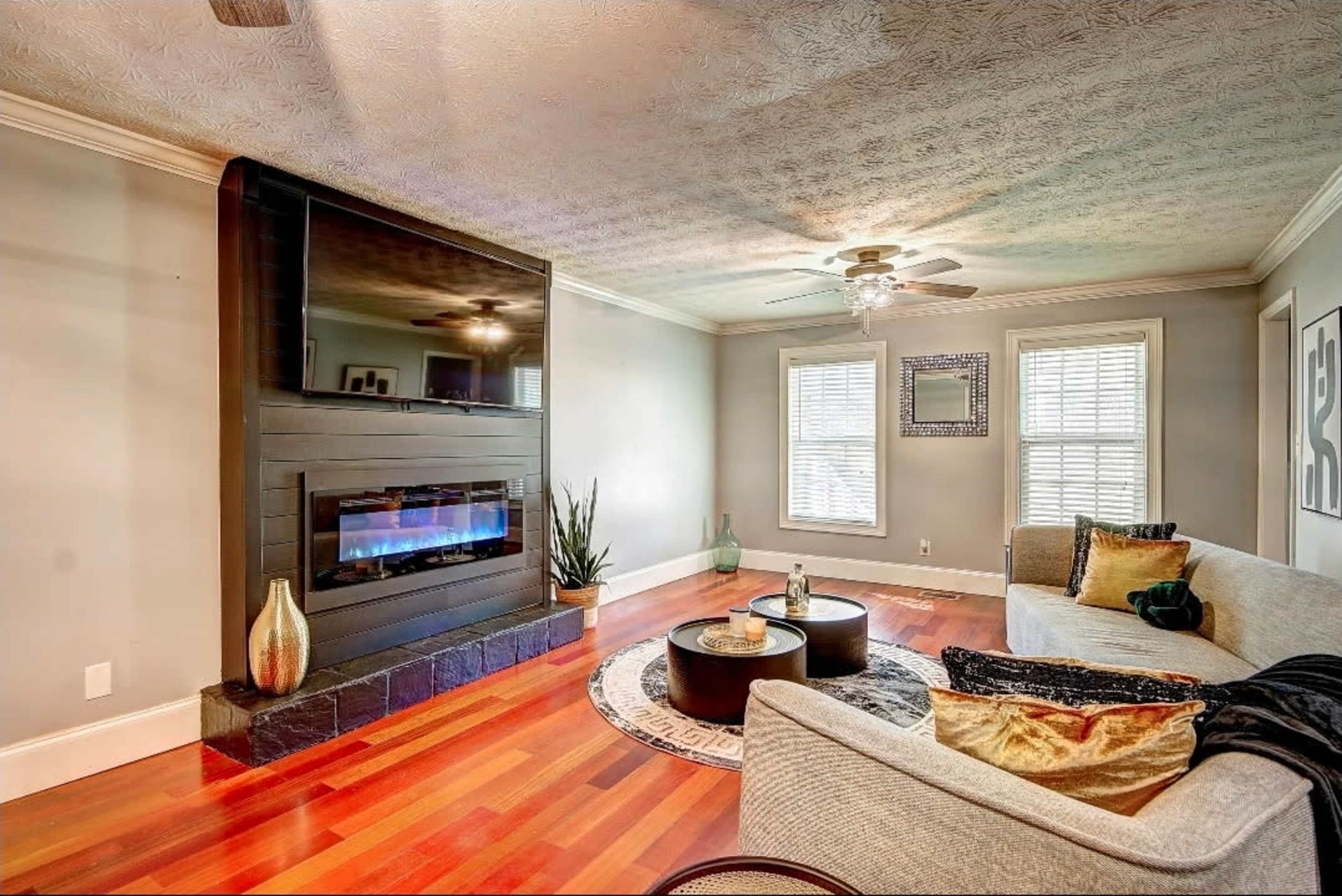 The image shows a modern living room featuring a wall-mounted TV above a sleek fireplace, with a light-colored sofa, a coffee table, and large windows allowing natural light to enter.