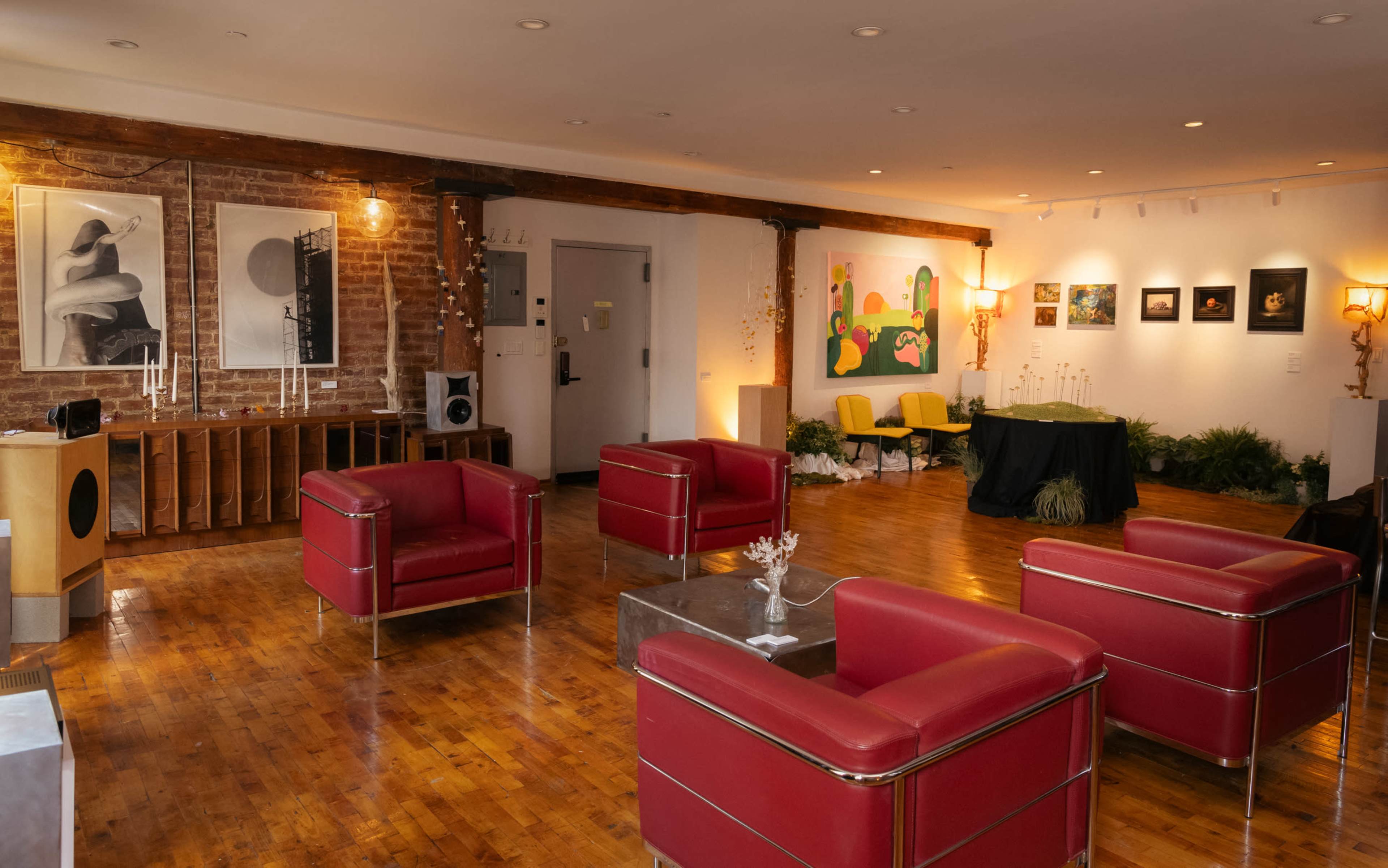 The scene depicts a spacious gallery with wooden floors, featuring red seating, artwork on the walls, and a display table near a collection of plants.