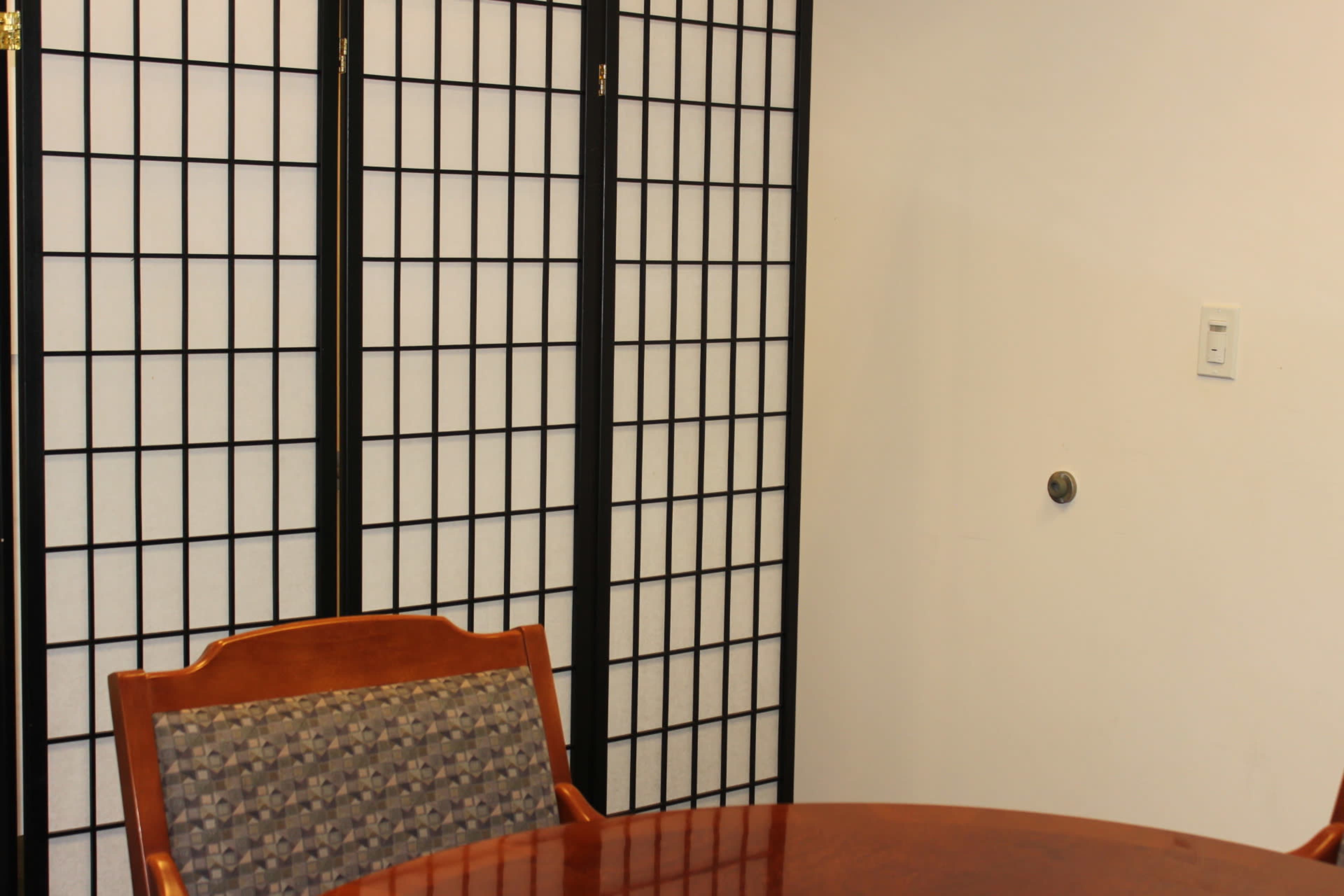 A wooden table with a patterned chair is set near a folding screen in a room with a plain white wall.