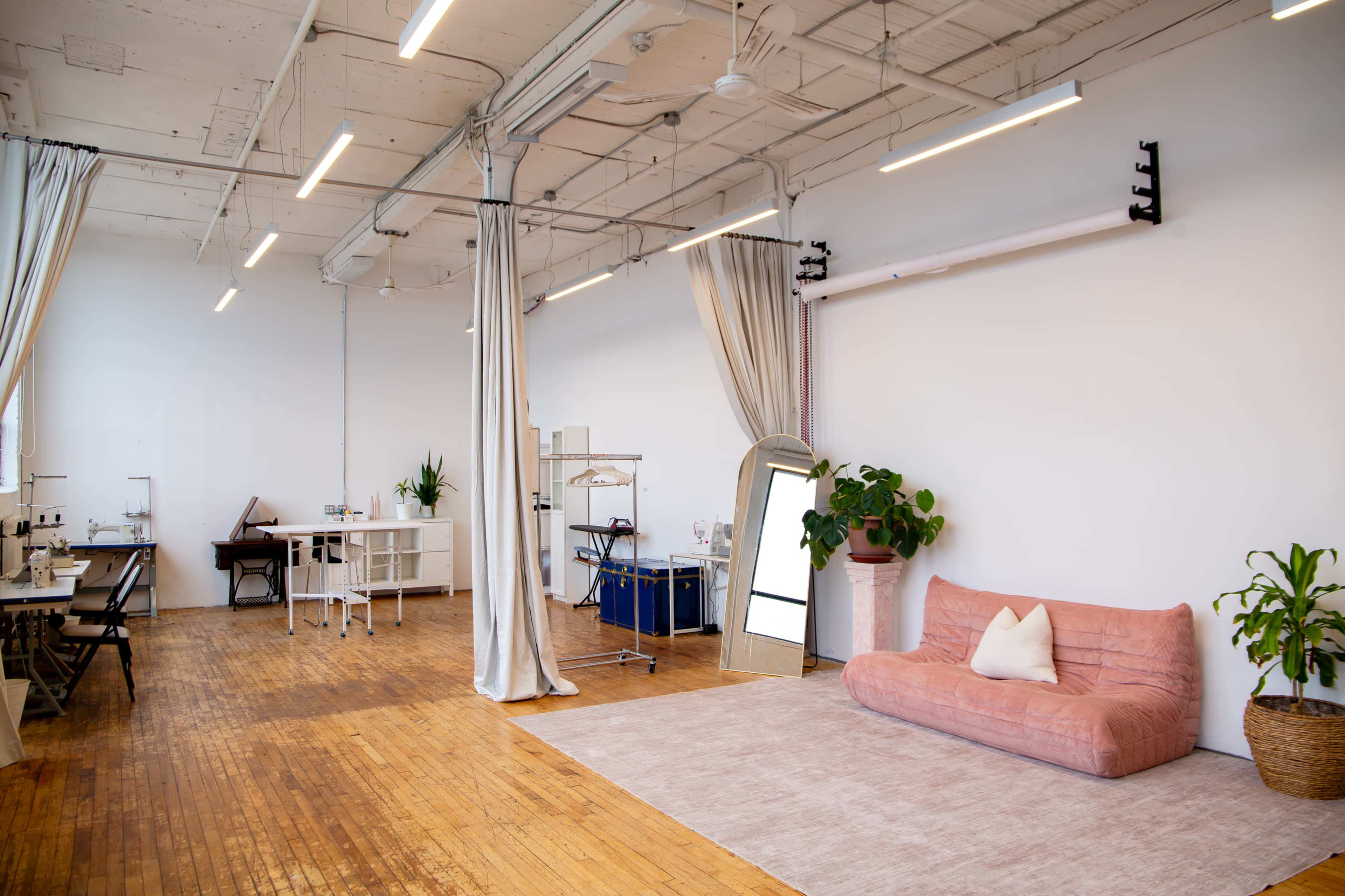 The image shows a spacious, well-lit studio with wooden flooring, featuring a pink sofa, plants, and a large mirror, along with workstations for sewing and a clothing rack.