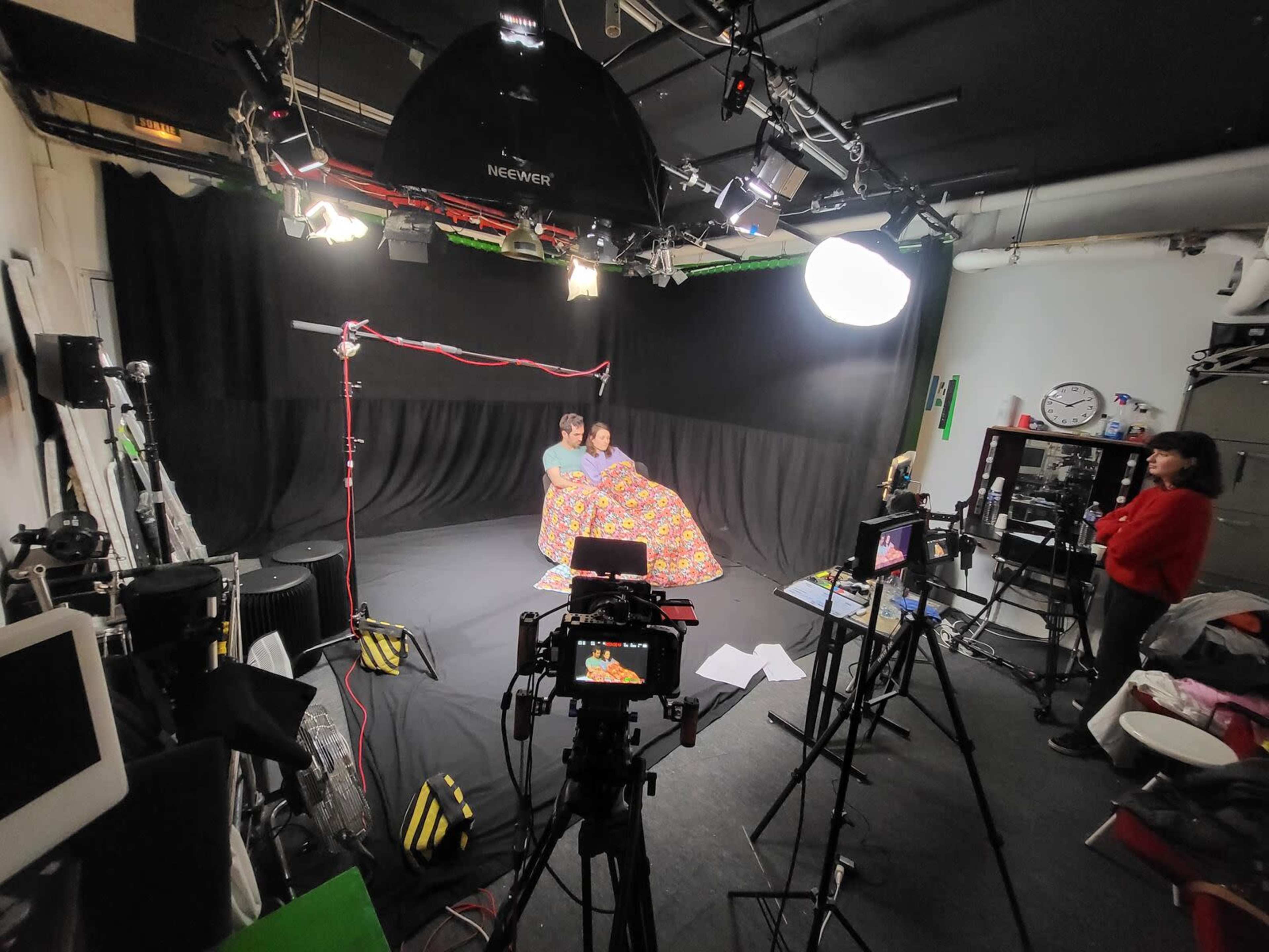 Two people sit under a colorful blanket in a photography studio surrounded by cameras and lighting equipment.