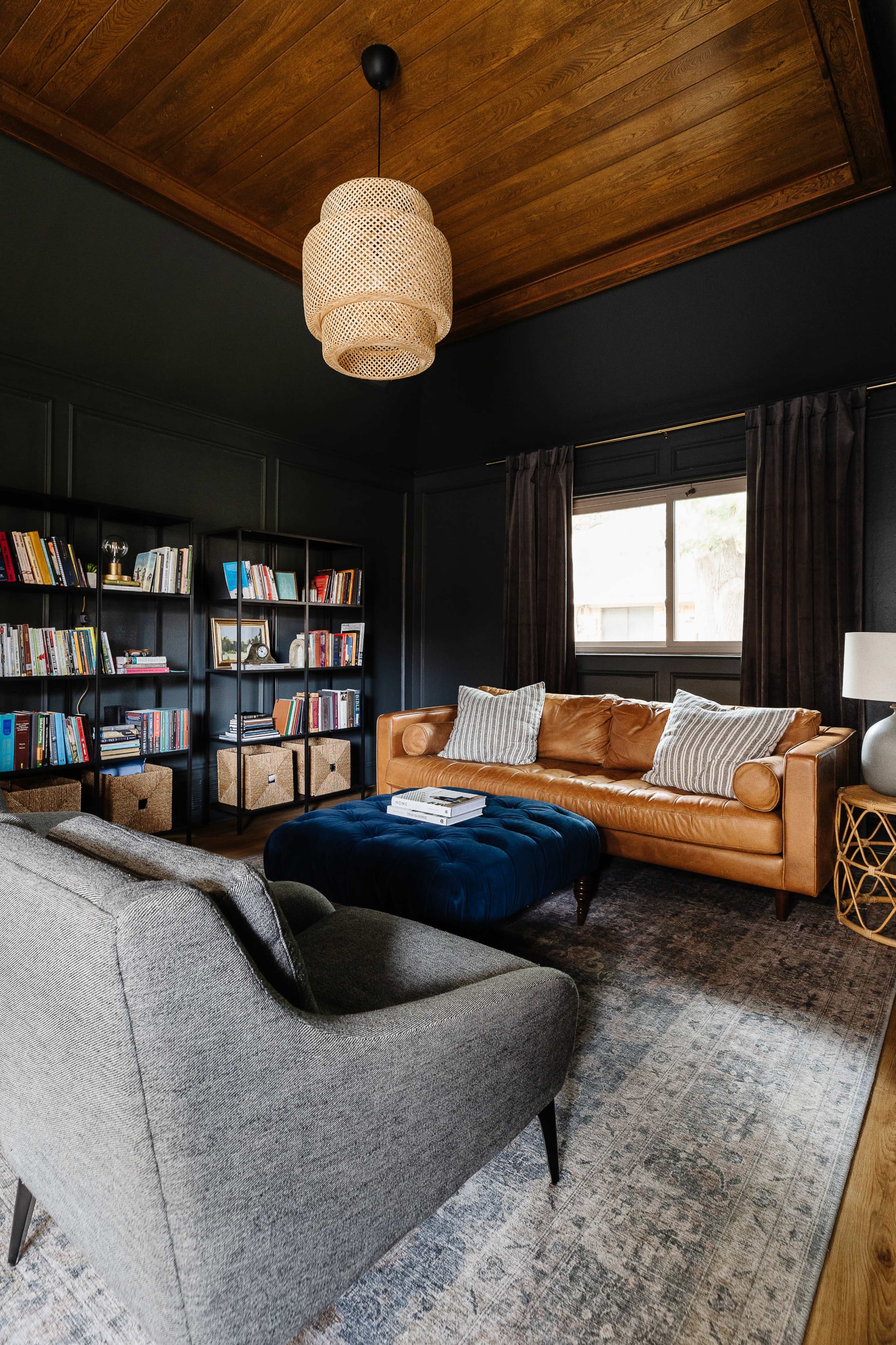 A contemporary living room featuring a brown leather sofa, a gray armchair, a dark wooden ceiling, and bookshelves filled with books.