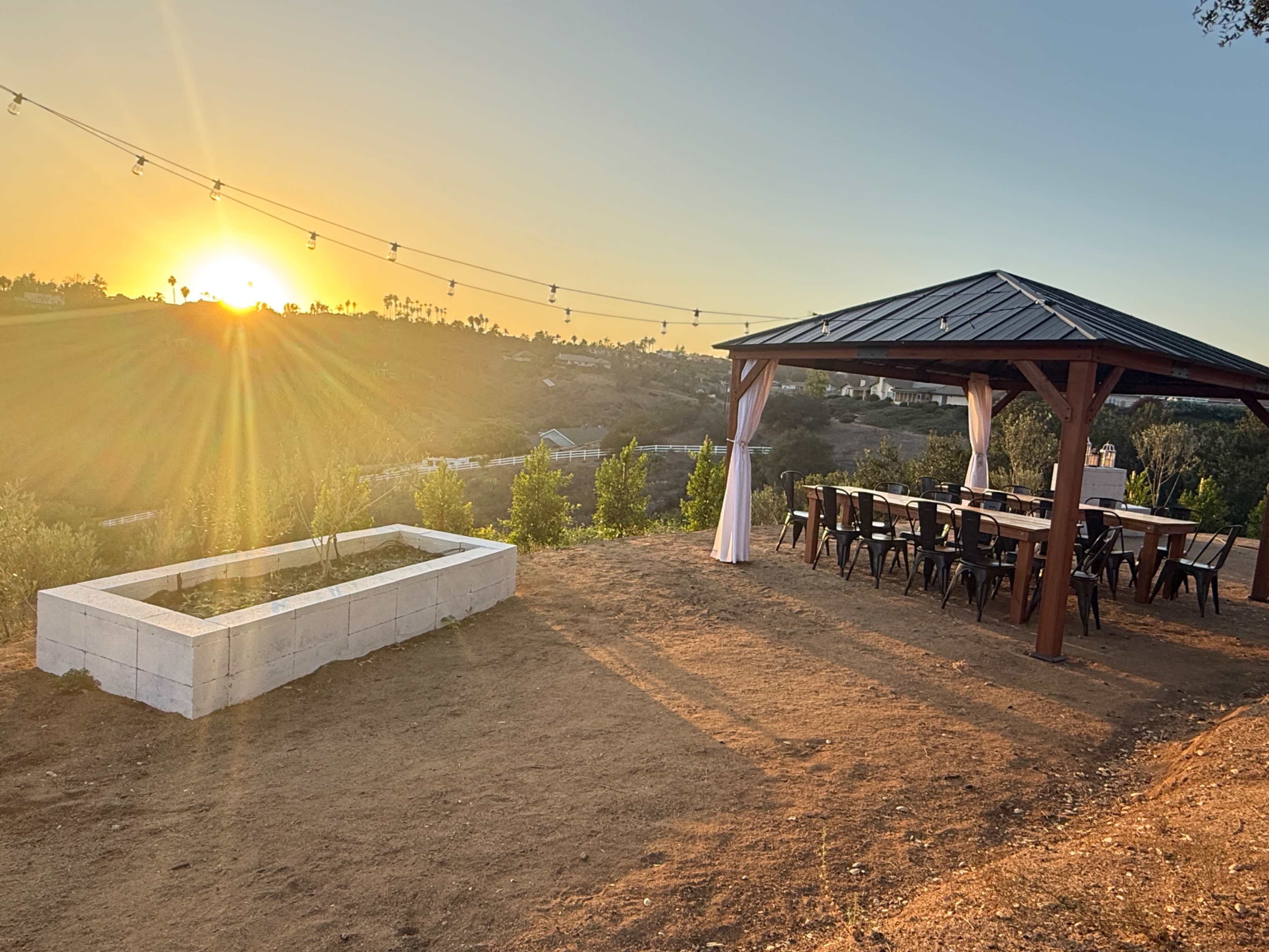 A wooden gazebo with a long table and chairs is set against a sunset backdrop, surrounded by greenery and a small garden bed.