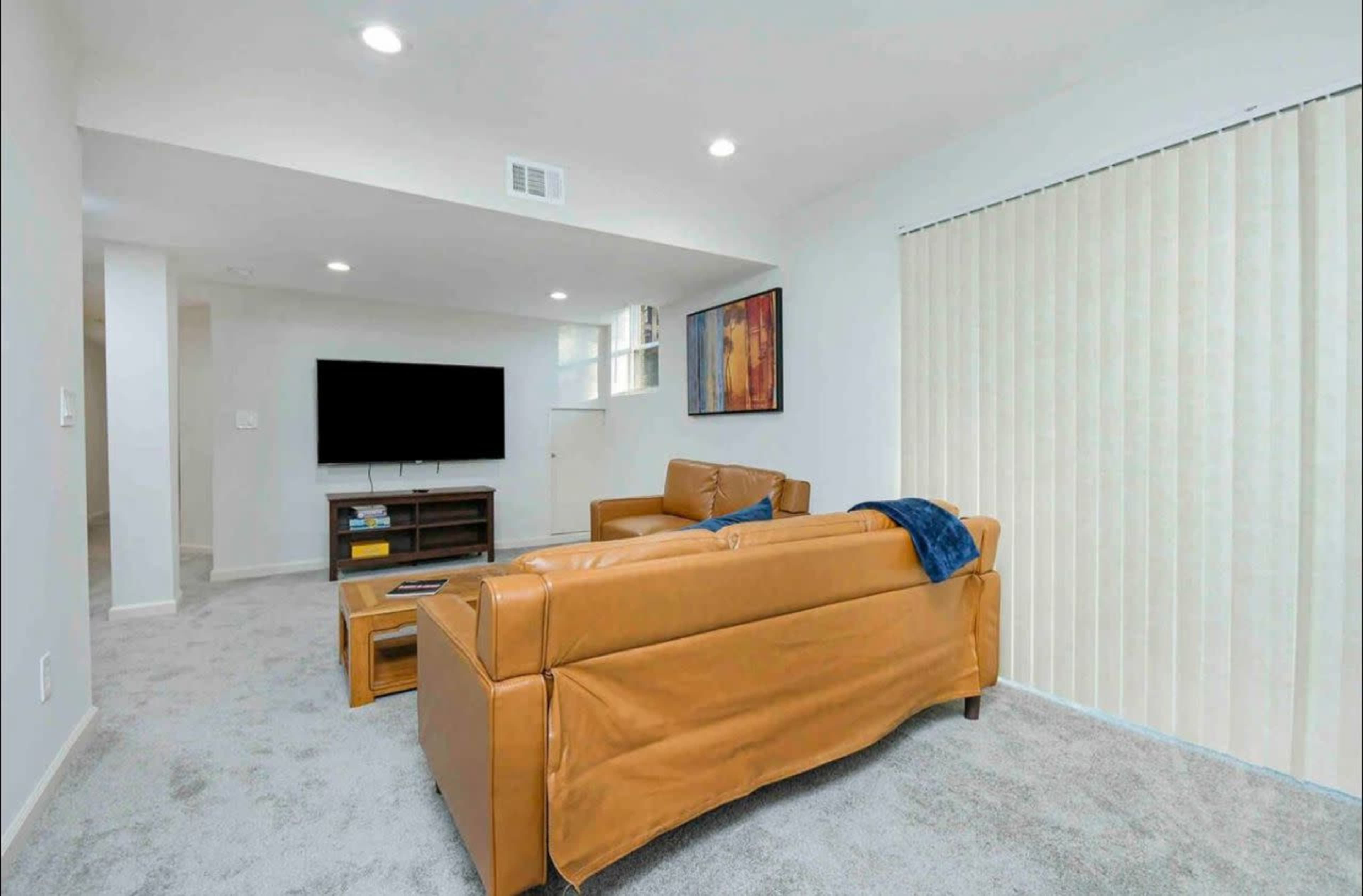 The image shows a modern living room with a light grey carpet, a brown leather sofa, a wooden media console, and a large television mounted on the wall.