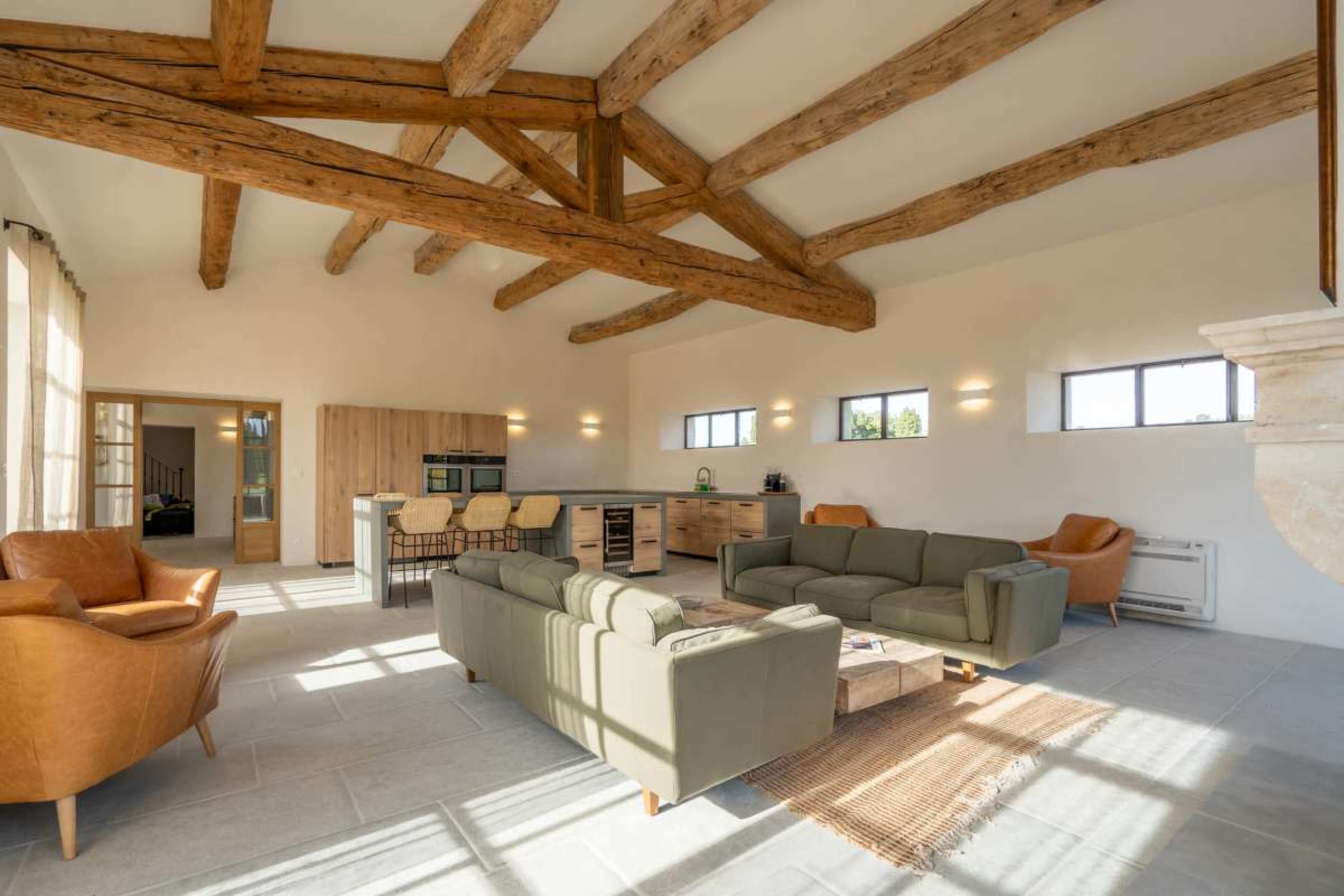 The image shows a spacious living area with exposed wooden beams, light-colored walls, and a mix of modern furniture including green sofas and brown armchairs.