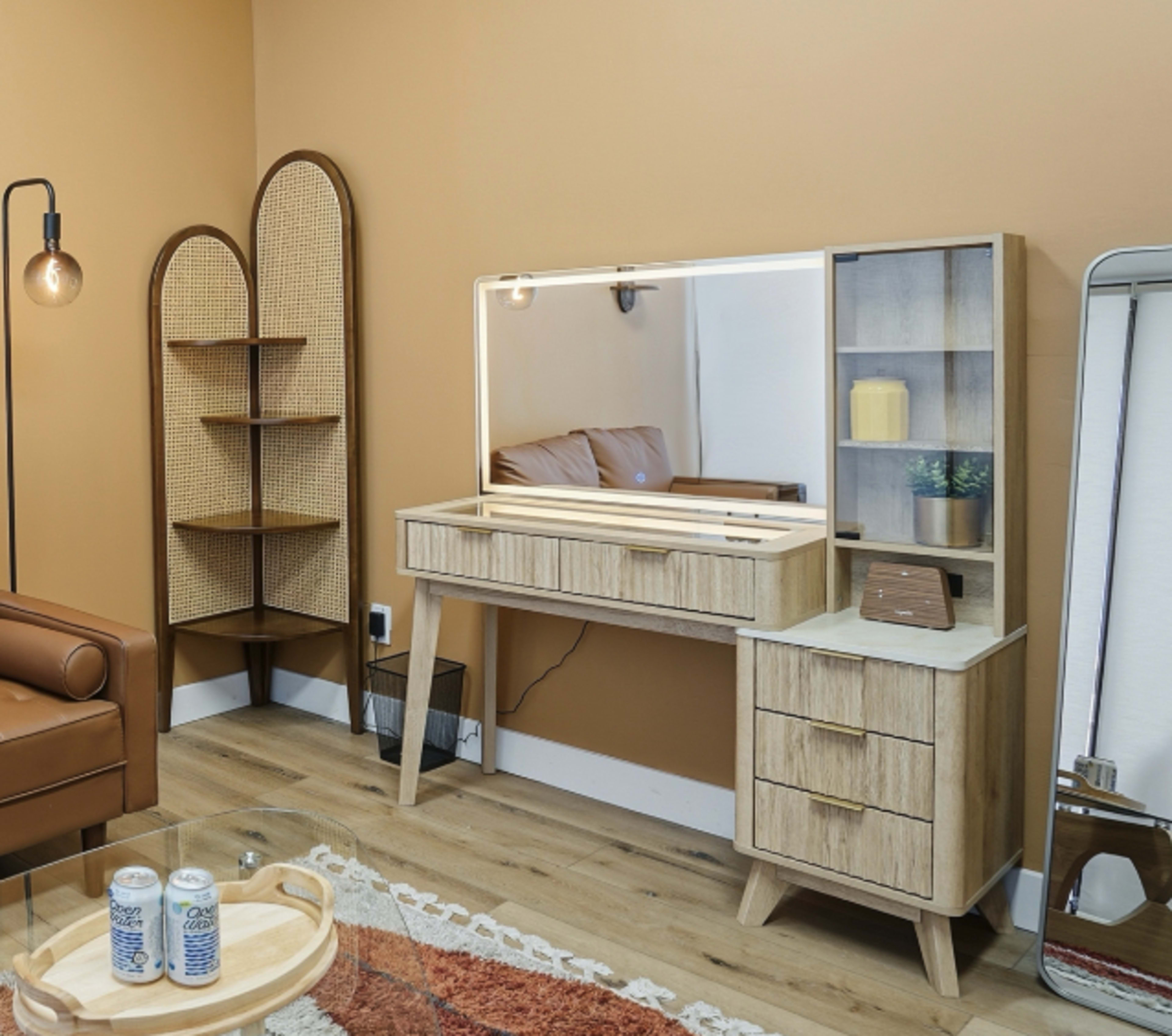 The image shows a modern interior space featuring a wooden vanity with a large mirror and drawer units, a tall shelf unit, a brown leather couch, and a glass coffee table.