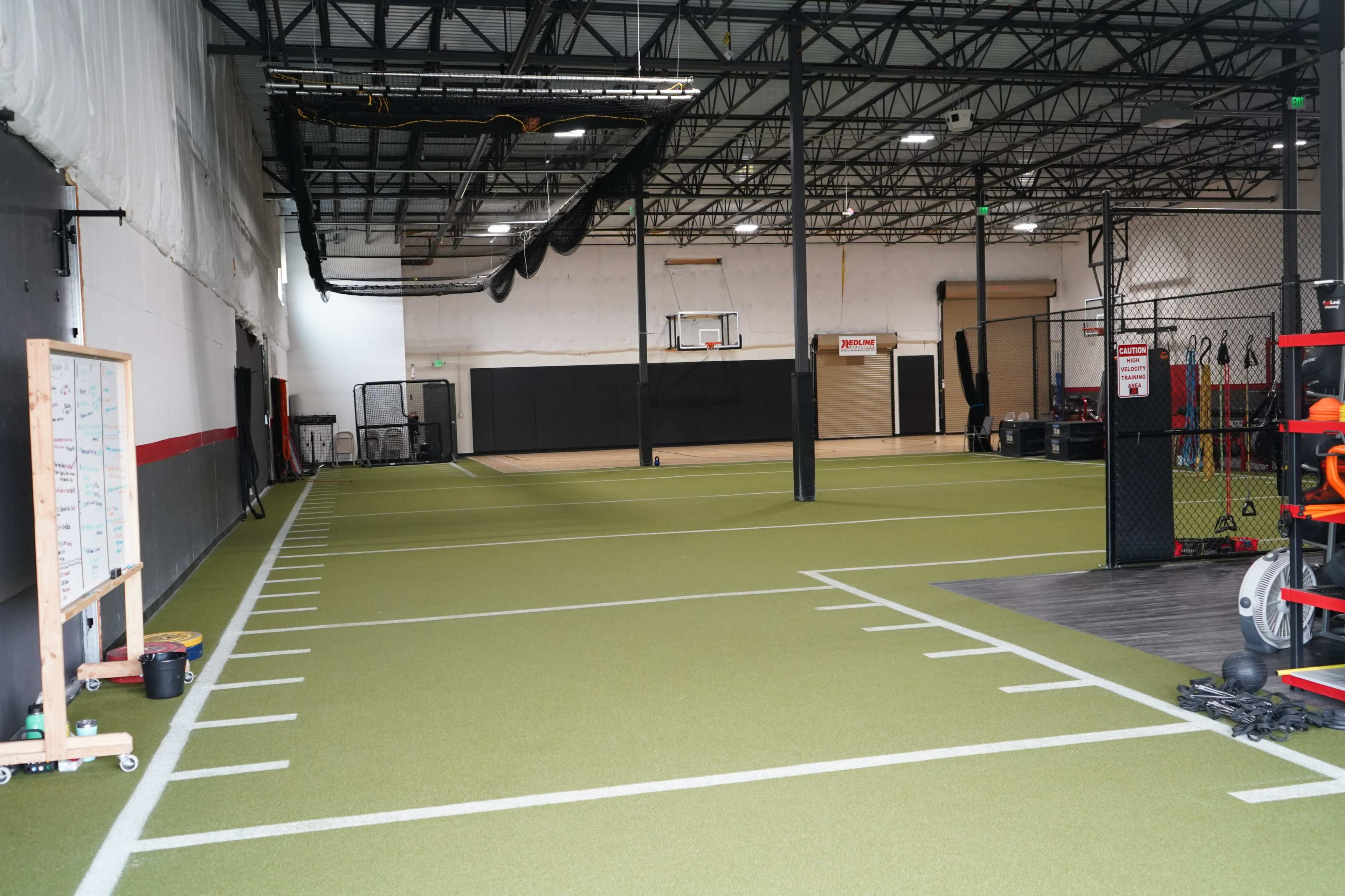 The image shows an indoor training facility with a green turf area, a basketball hoop in the background, and various fitness equipment along one side.