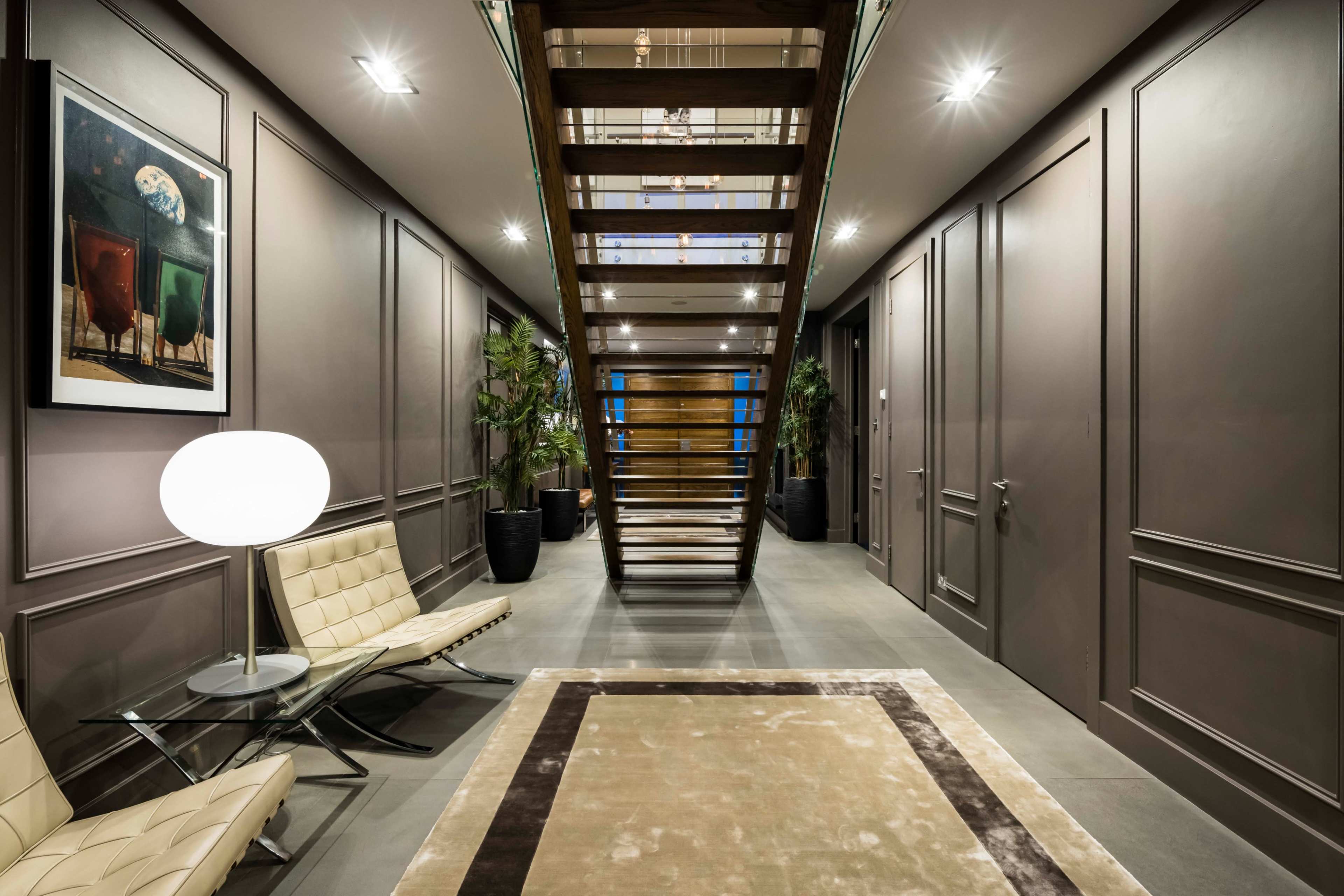 The image shows a modern interior hallway with a staircase at the center, framed by contemporary furniture and plants.