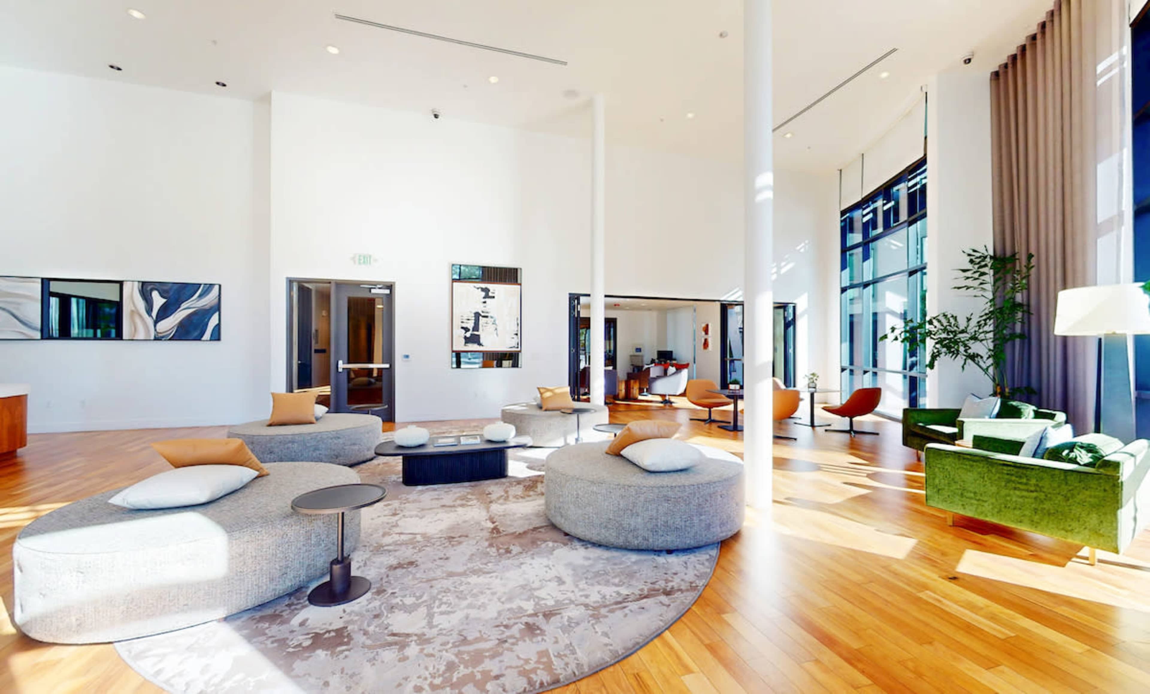 The image shows a spacious, modern lounge area with circular seating, a decorative rug, and large windows allowing natural light to enter.