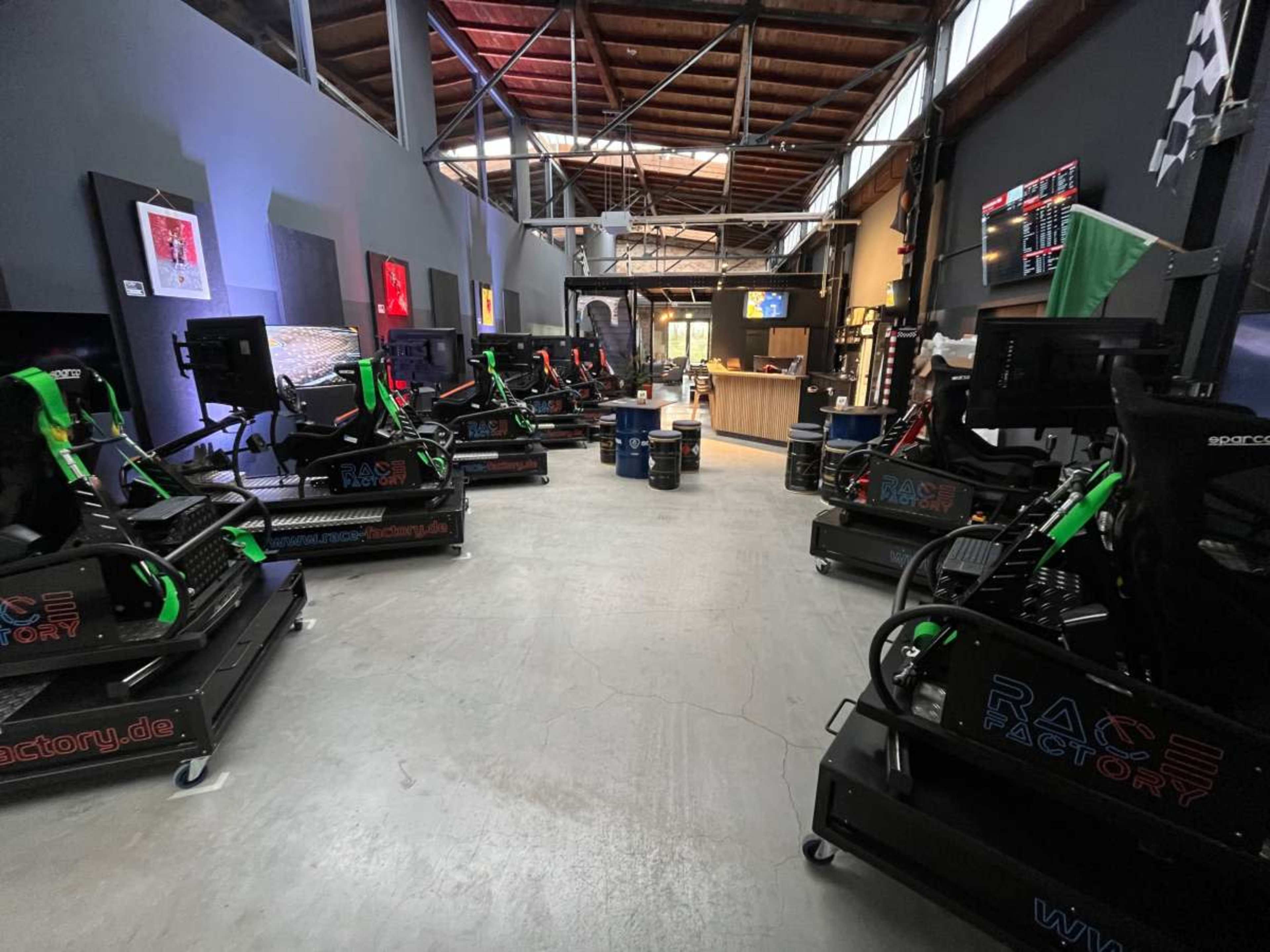 The image shows a spacious indoor venue featuring multiple racing simulators arranged in a row, with a counter and various decorations in the background.