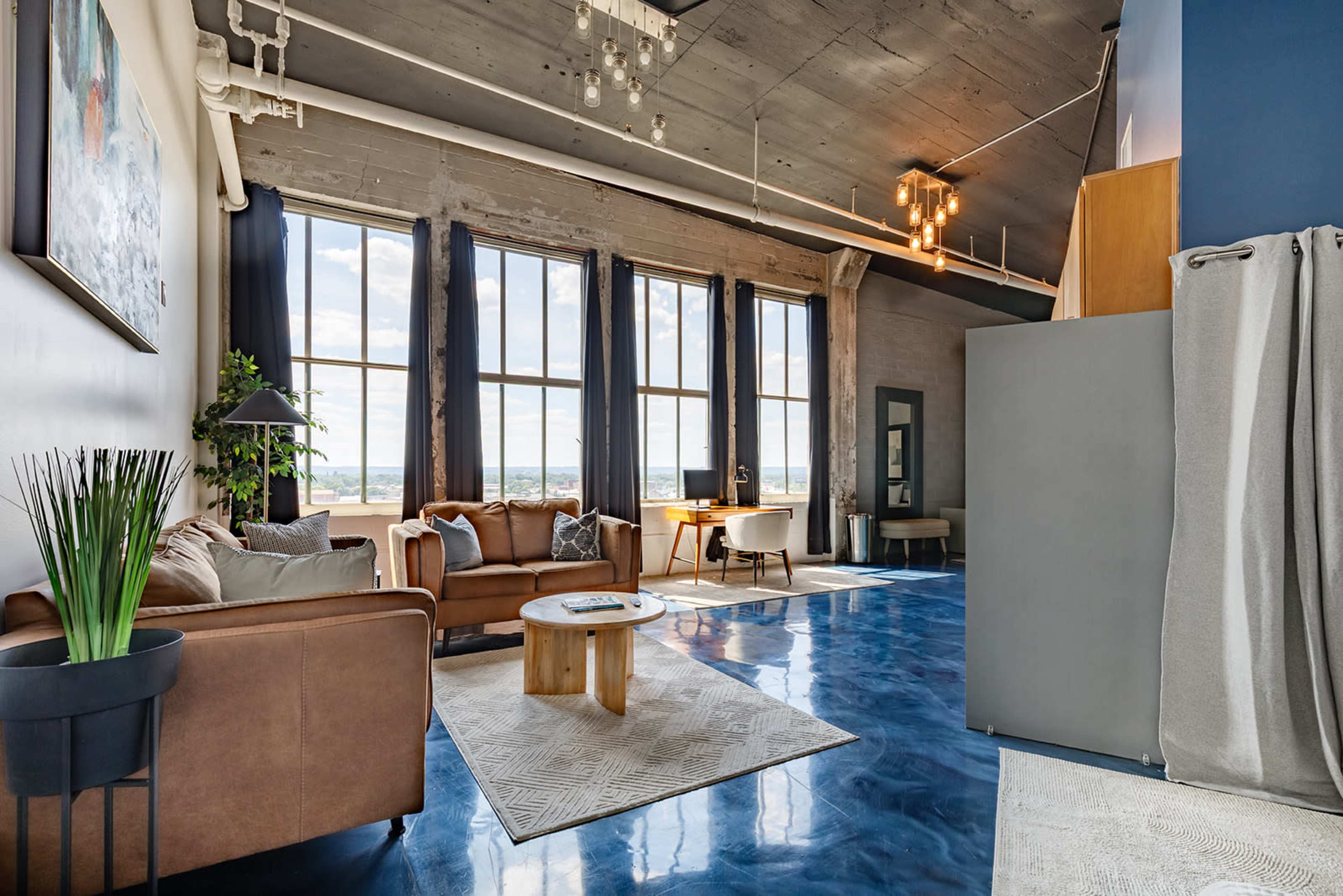 The image shows a modern living room with a brown sofa, a round wooden coffee table, and large windows allowing for ample natural light.