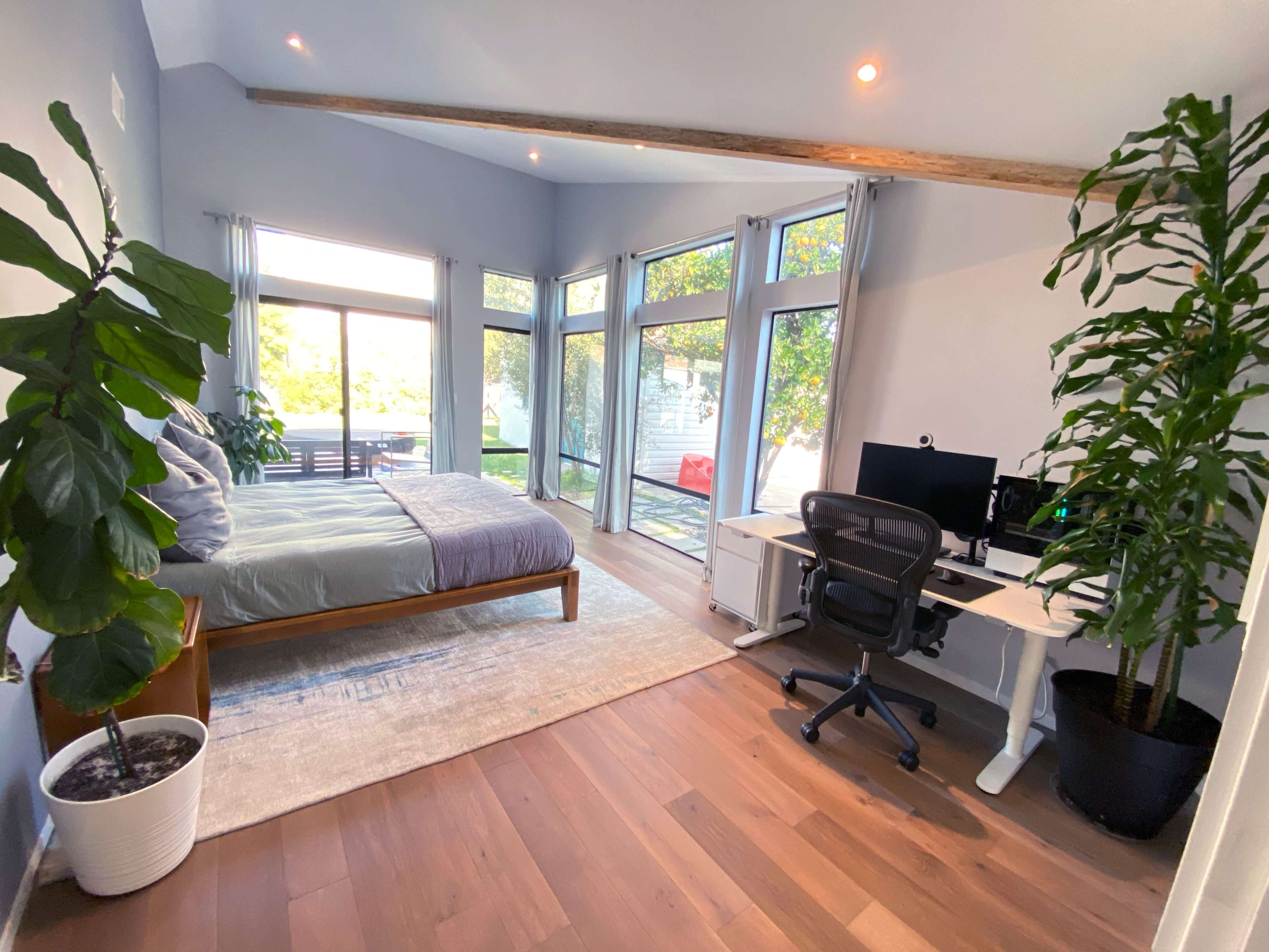 A spacious, well-lit bedroom featuring a bed, a desk with a computer, and large windows overlooking an outdoor area.