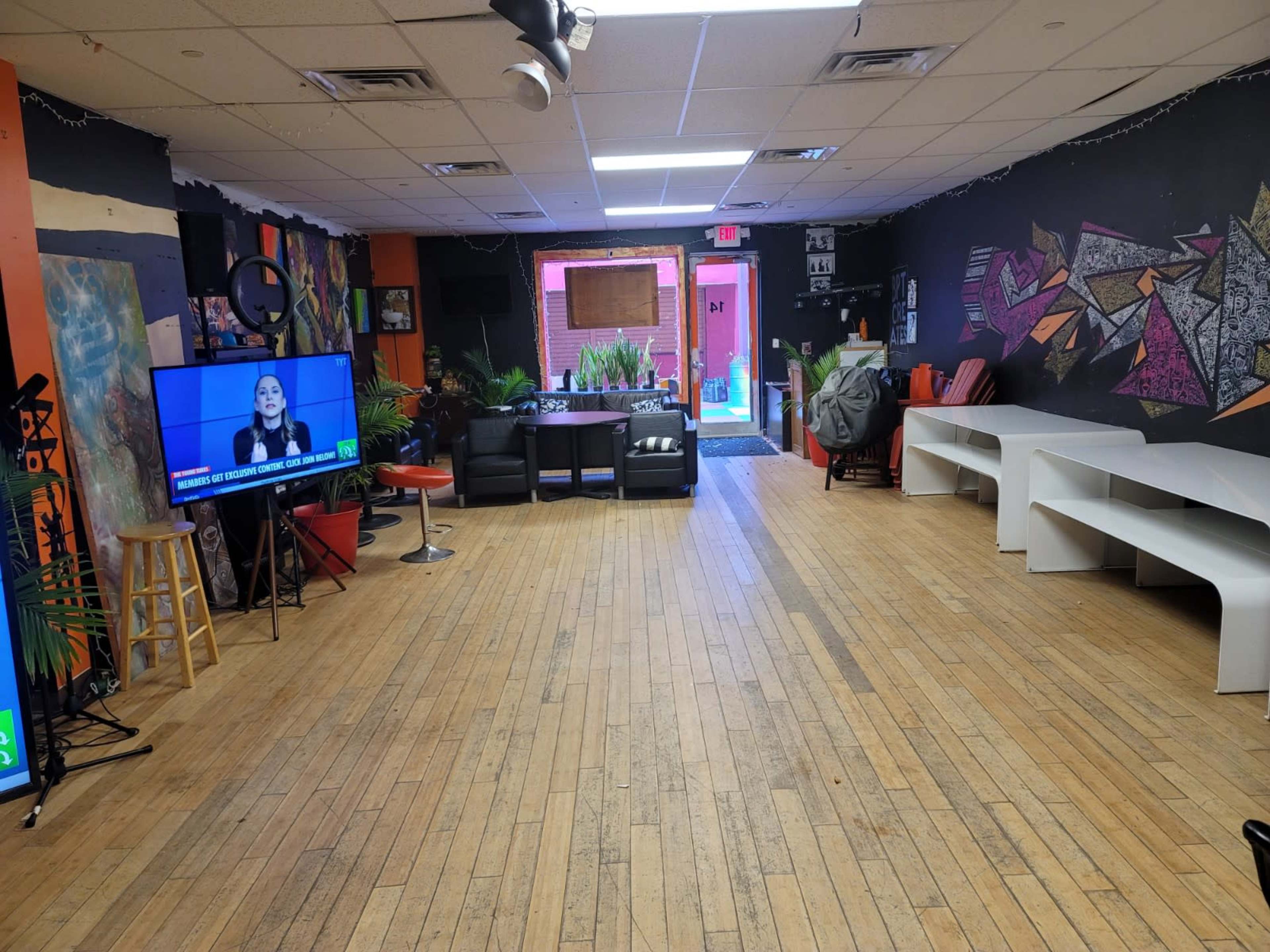 The image shows a spacious room with hardwood flooring, featuring a television displaying a program, a black sofa set, several white tables, and vibrant wall art.