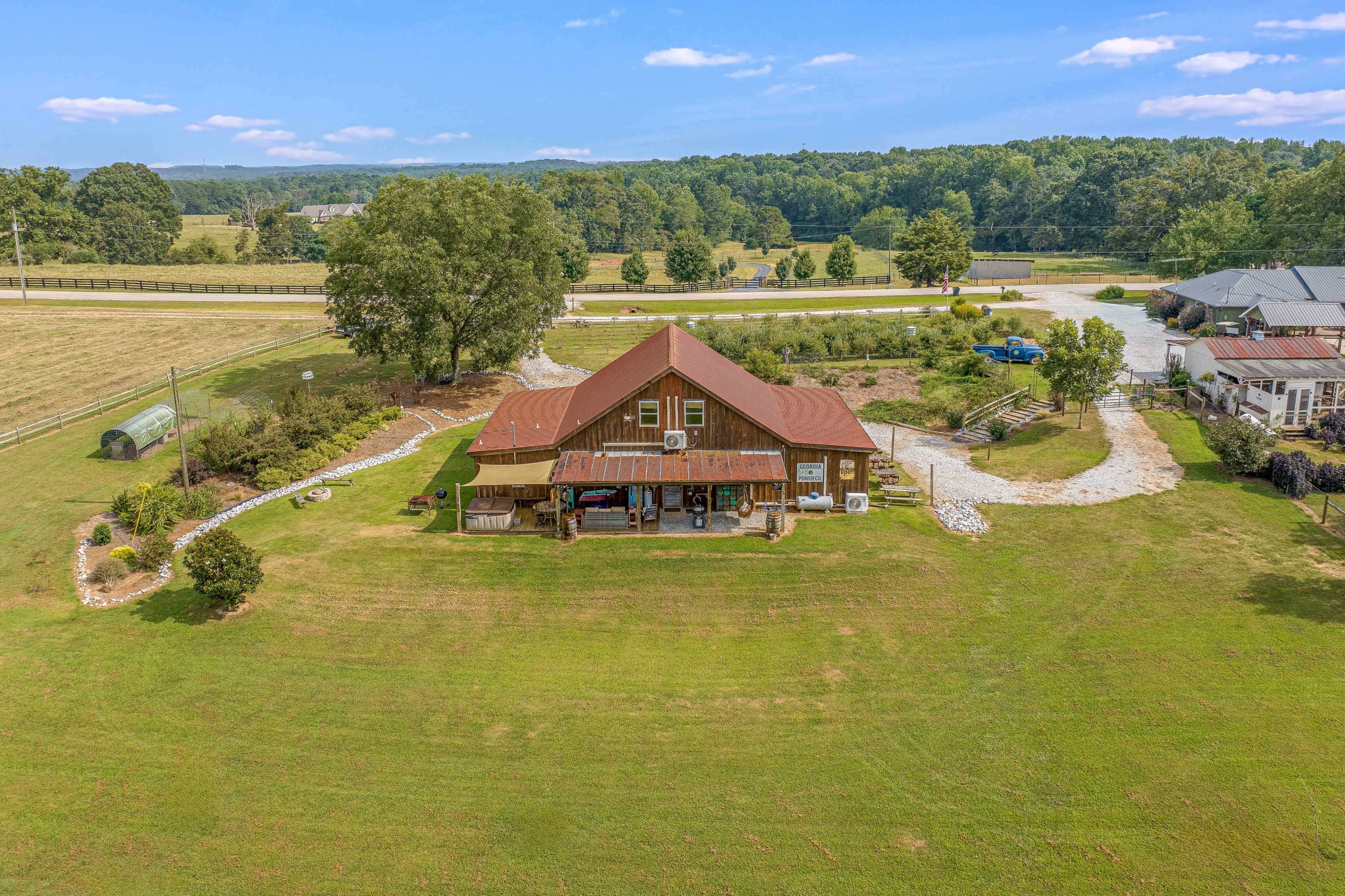 A large wooden farmhouse with a red metal roof is situated on a grassy area surrounded by trees and a gravel driveway.