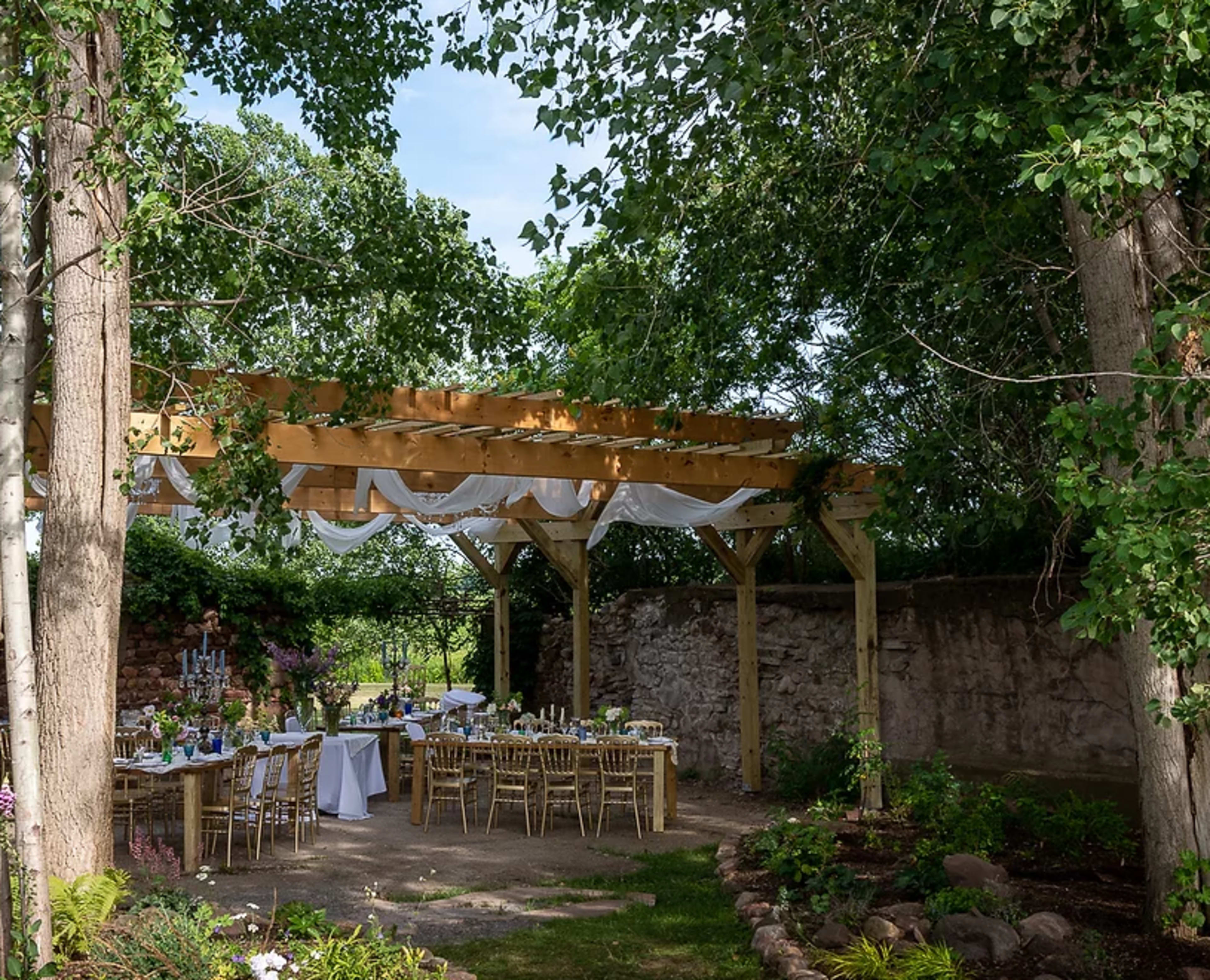 A wooden pergola draped with white fabric stands in a green garden, surrounded by trees and set with tables for dining.