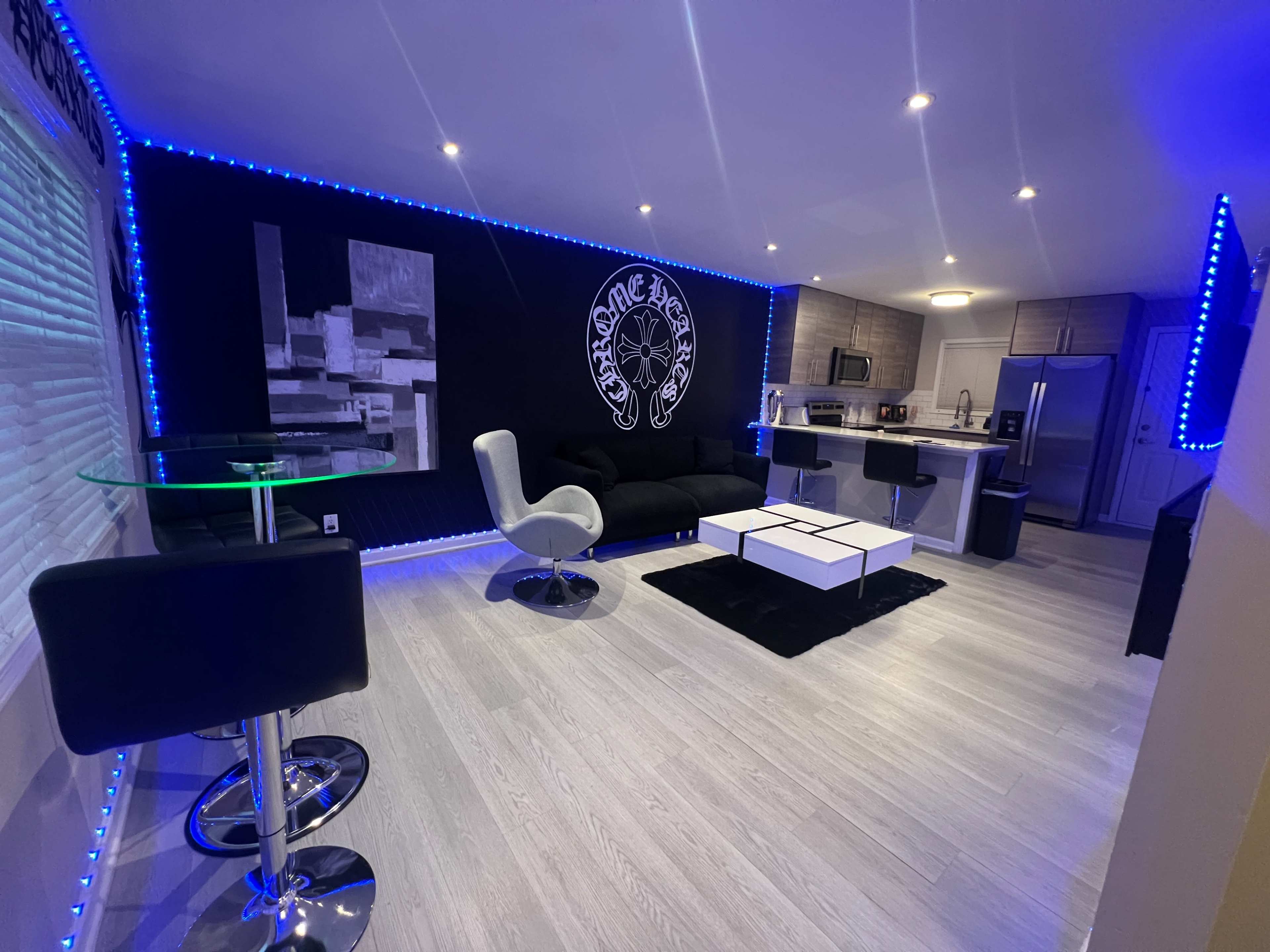 The image shows a modern, stylish living space with a black and white color scheme, featuring a black sofa, a glass dining table with black bar stools, and blue LED lighting along the walls.
