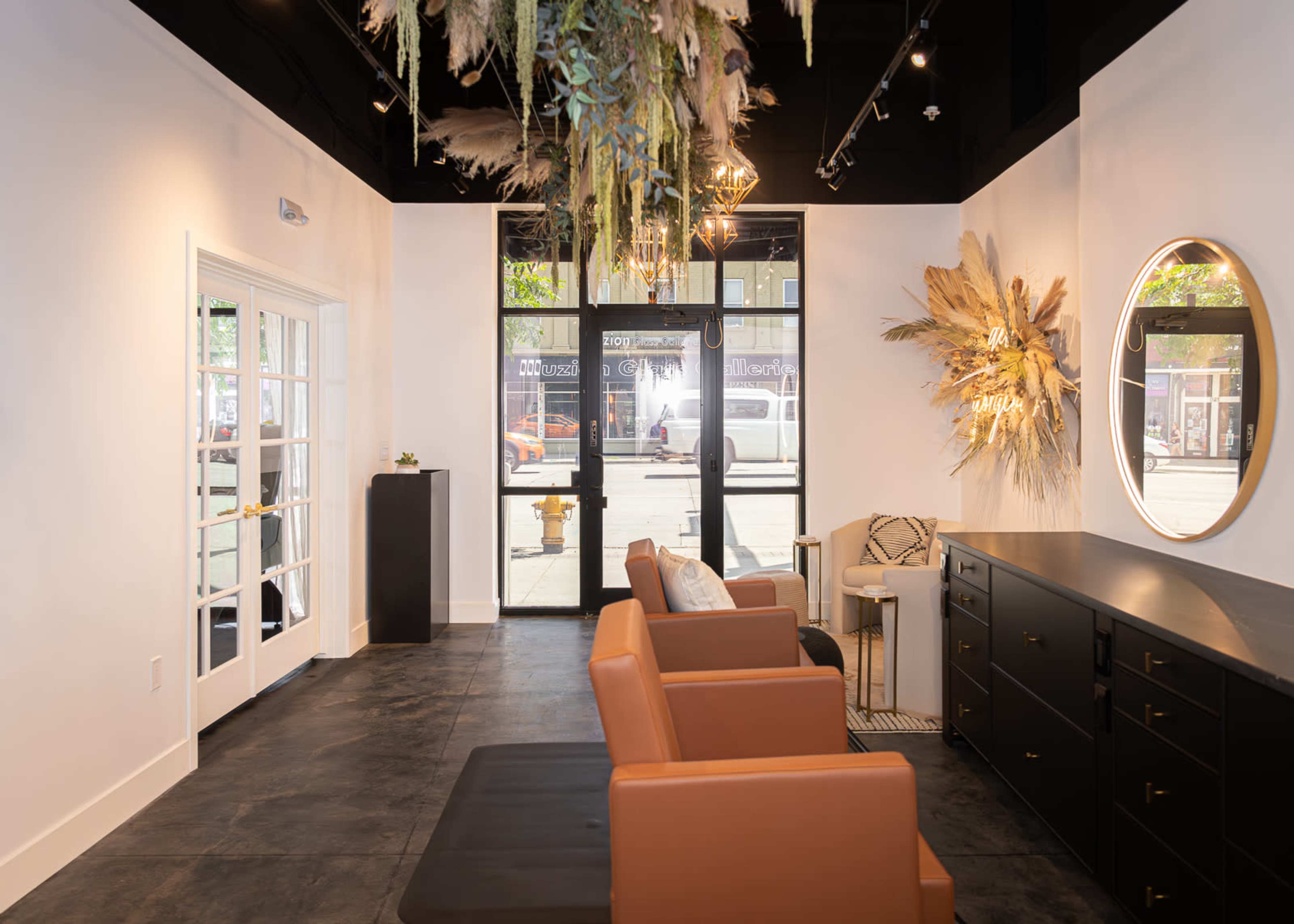 The image shows a modern interior space featuring brown leather seating, a dark cabinet, and decorative wall elements, with large windows allowing natural light to enter.
