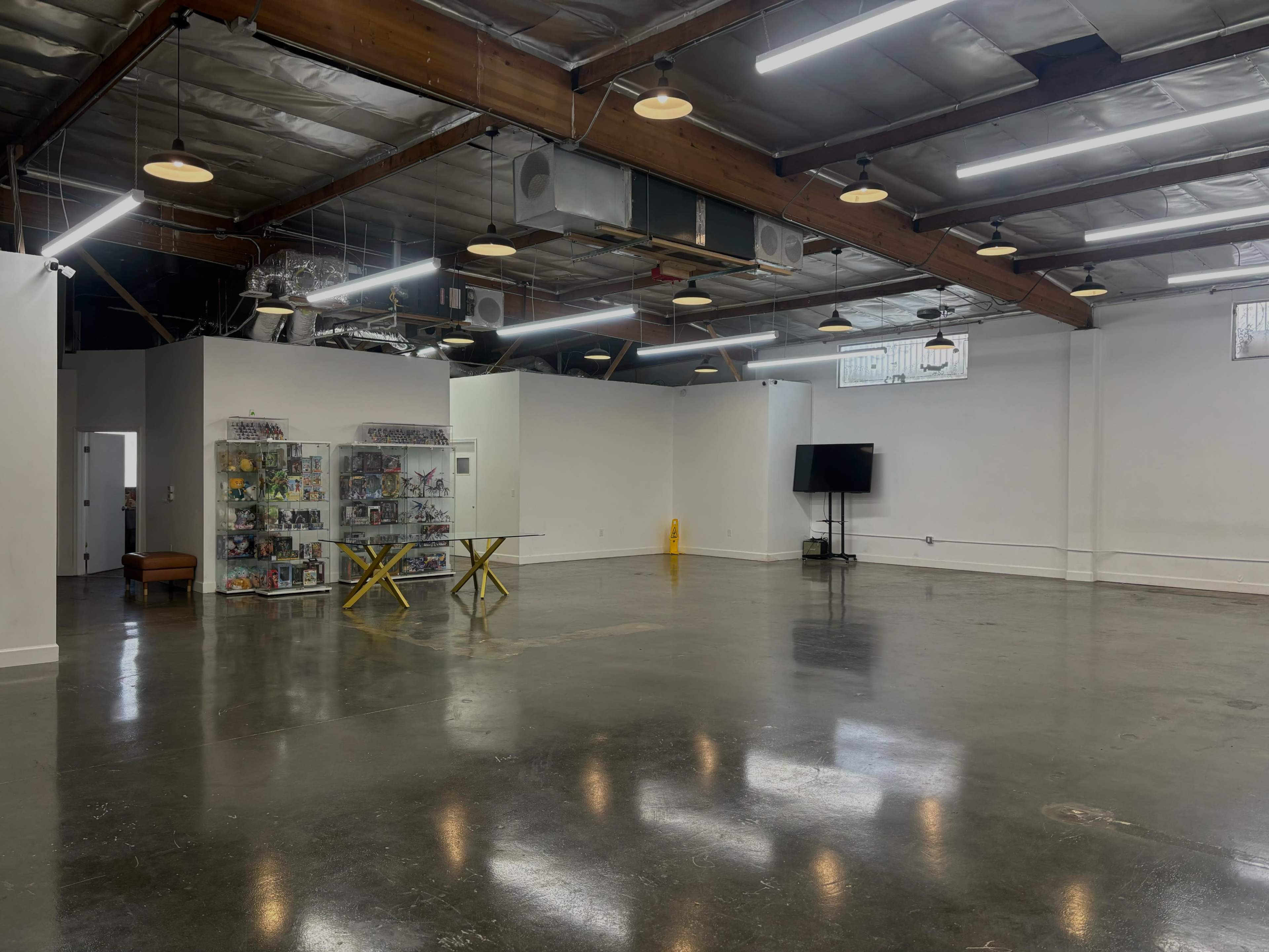 The image shows a spacious, empty room with polished concrete floors, overhead lights, a display shelf filled with various items, and a television mounted on a wall.