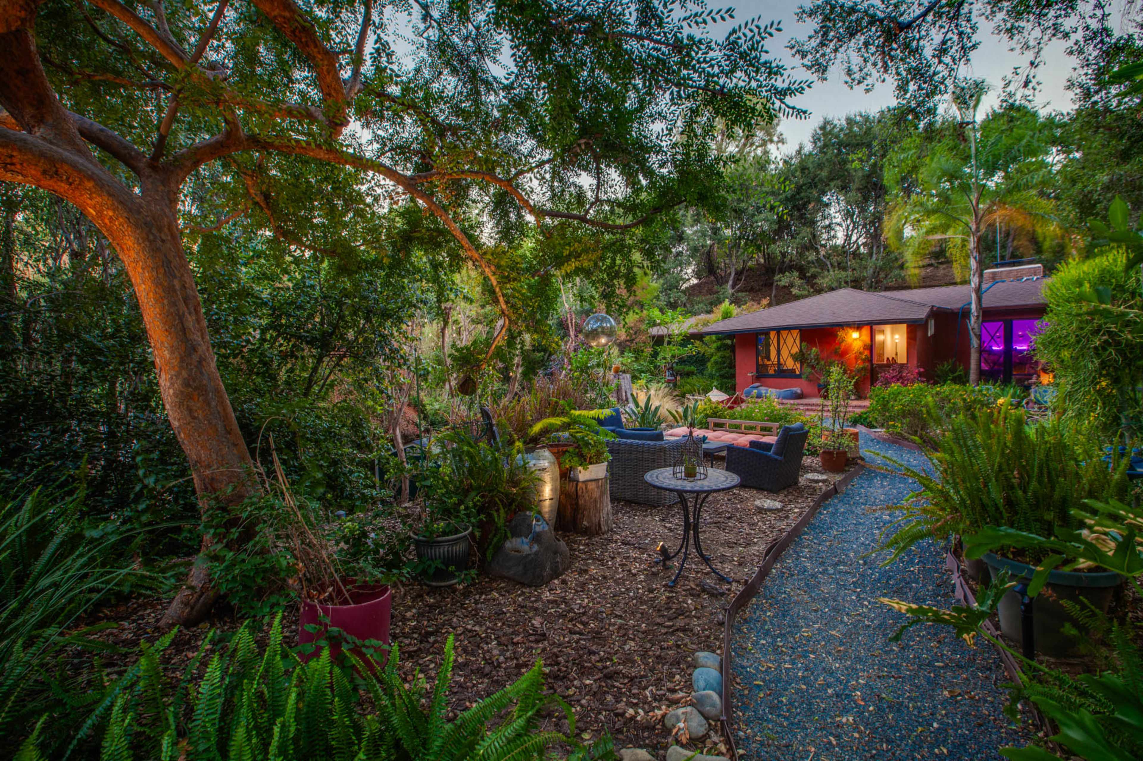 A serene garden scene features a winding pathway surrounded by lush greenery, leading to a cozy seating area near a house illuminated by soft lights.