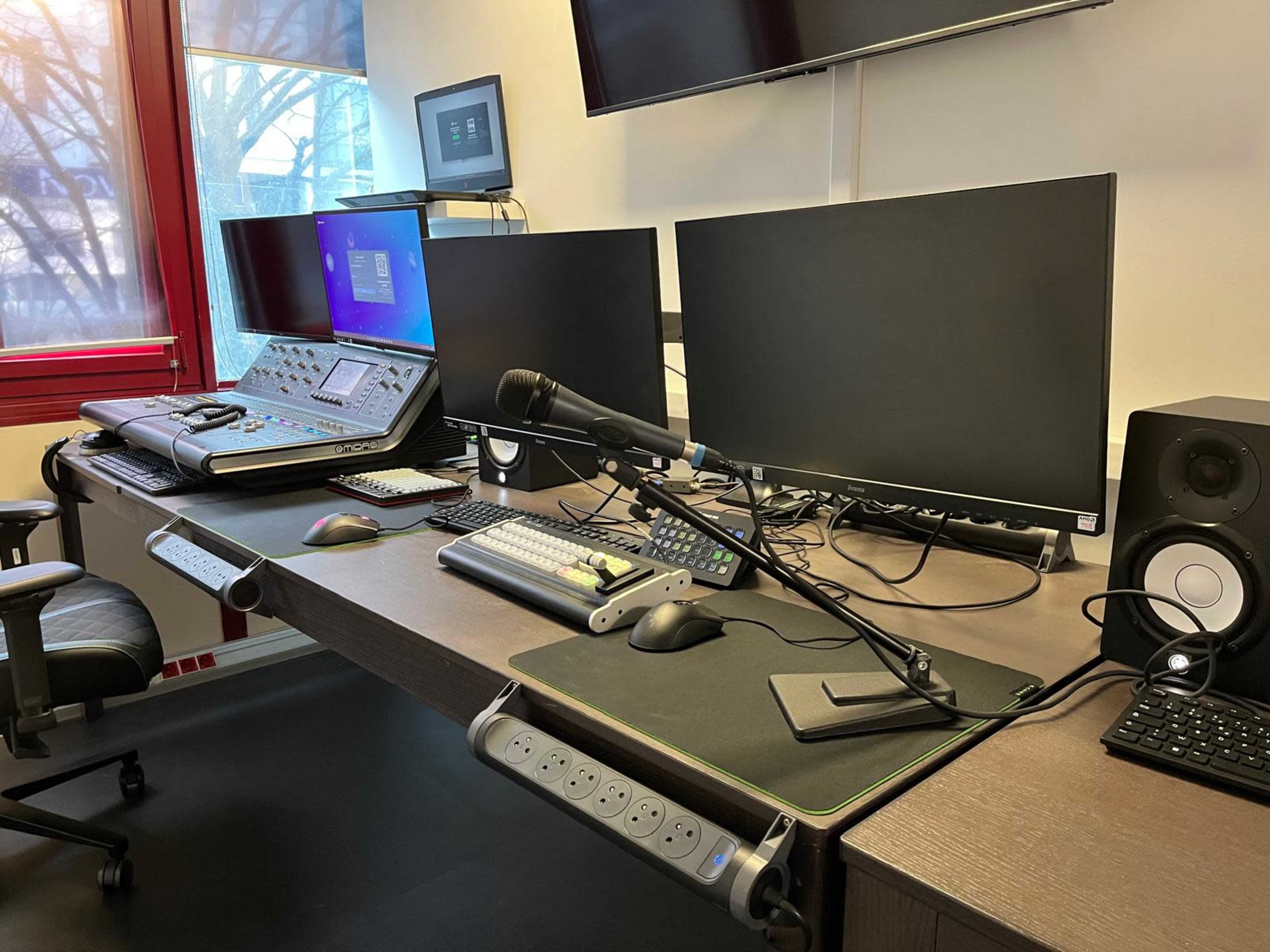 The image shows a modern workstation with two large monitors, a microphone, a control panel, and audio equipment arranged neatly on a wooden desk.
