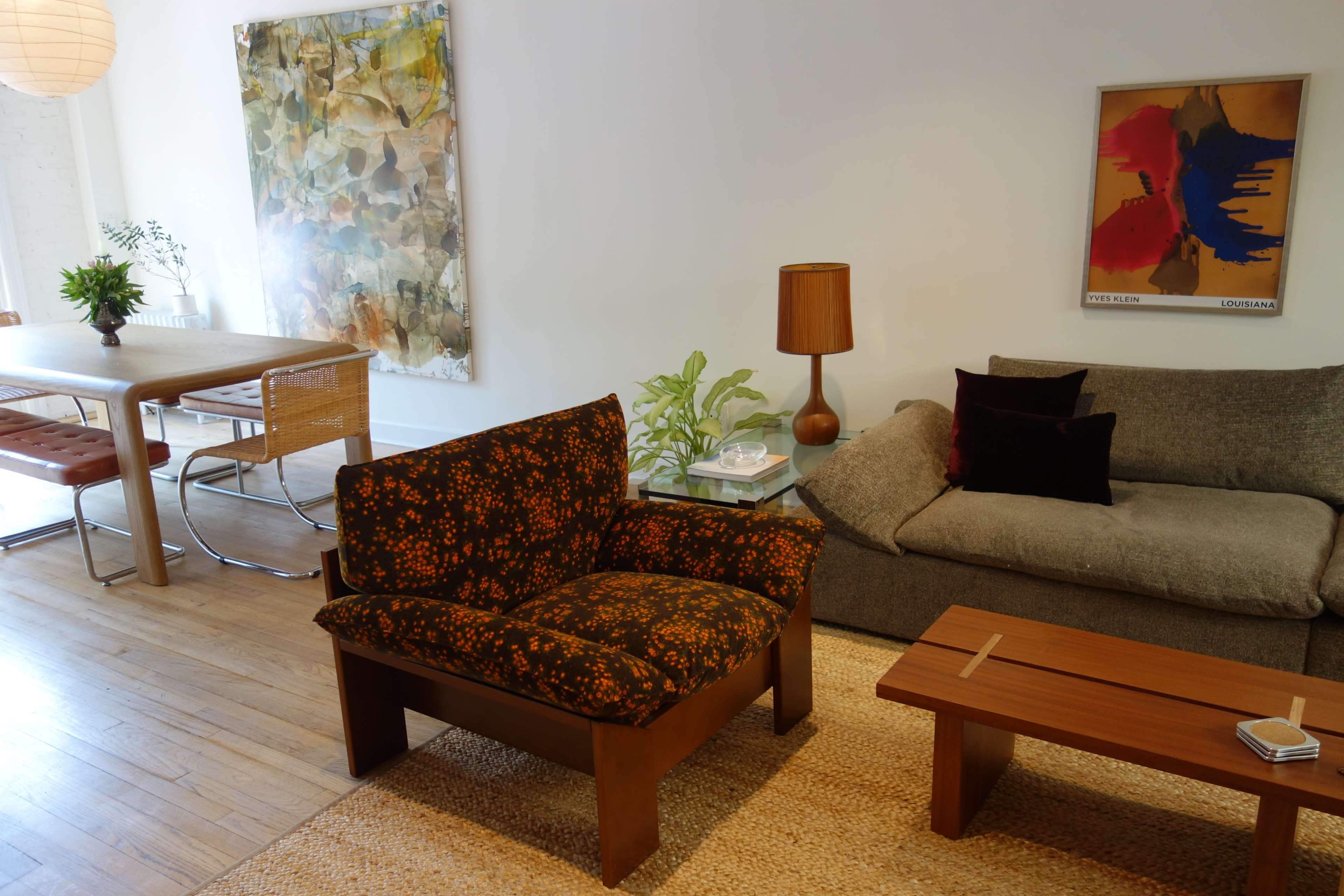 The image shows a modern living space featuring a patterned armchair, a gray sofa, a wooden coffee table, and a dining area with a glass-top table and metal chairs.