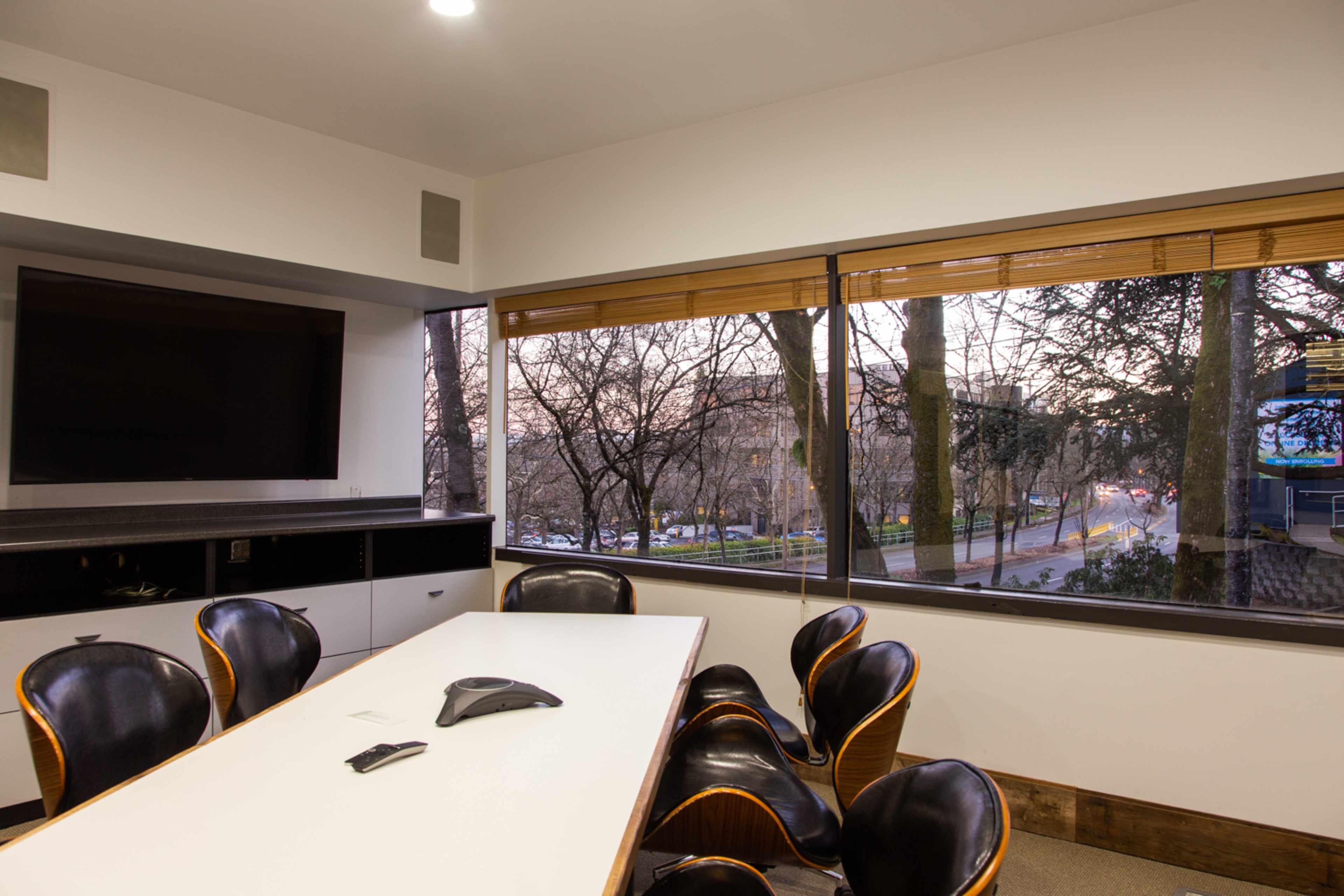 A conference room features a long table with black chairs and a large window overlooking a tree-lined street.