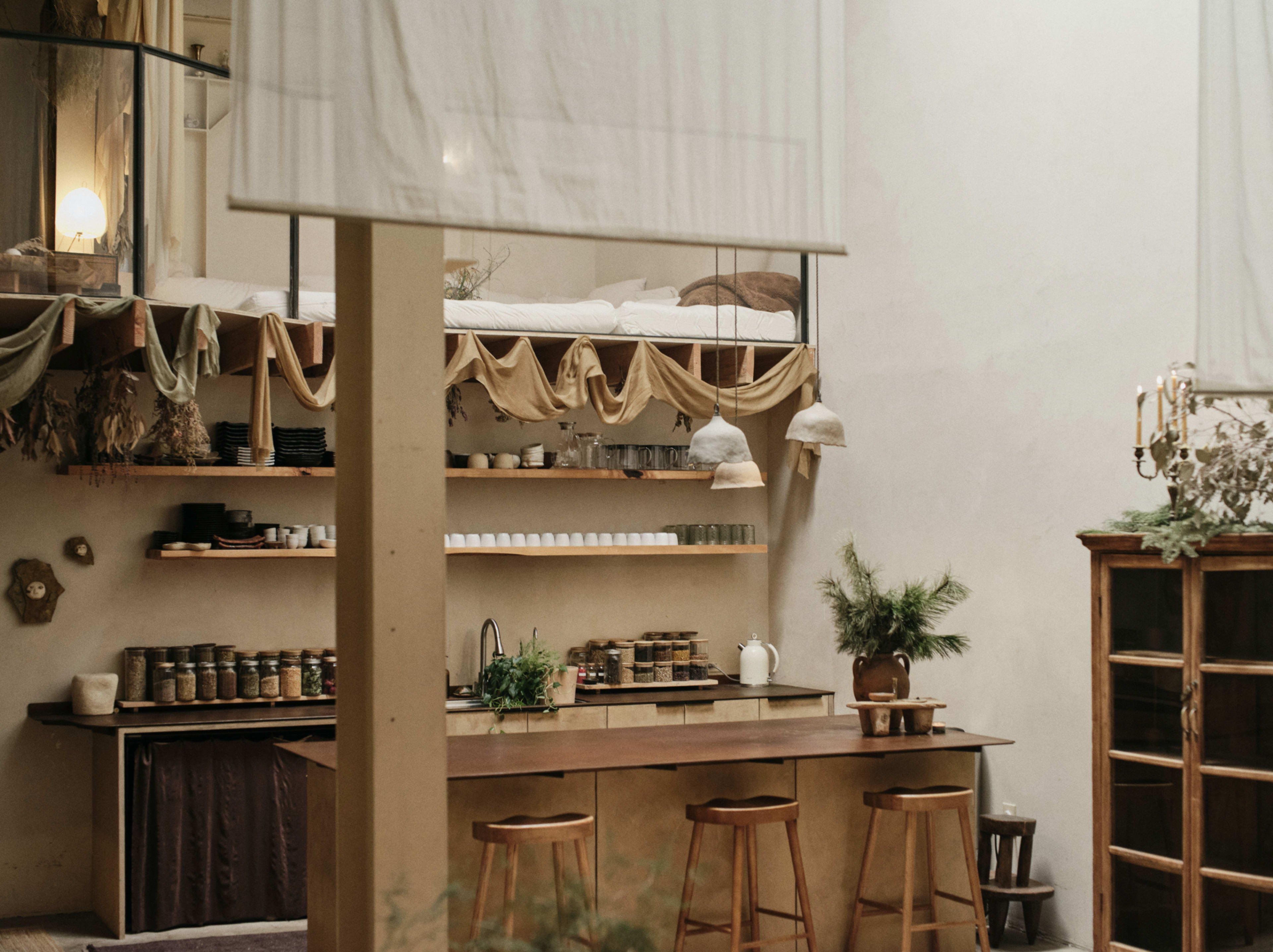 The image shows a minimalistic kitchen with wooden shelves, a central island with stools, and natural decor elements, all set in a spacious, well-lit room.