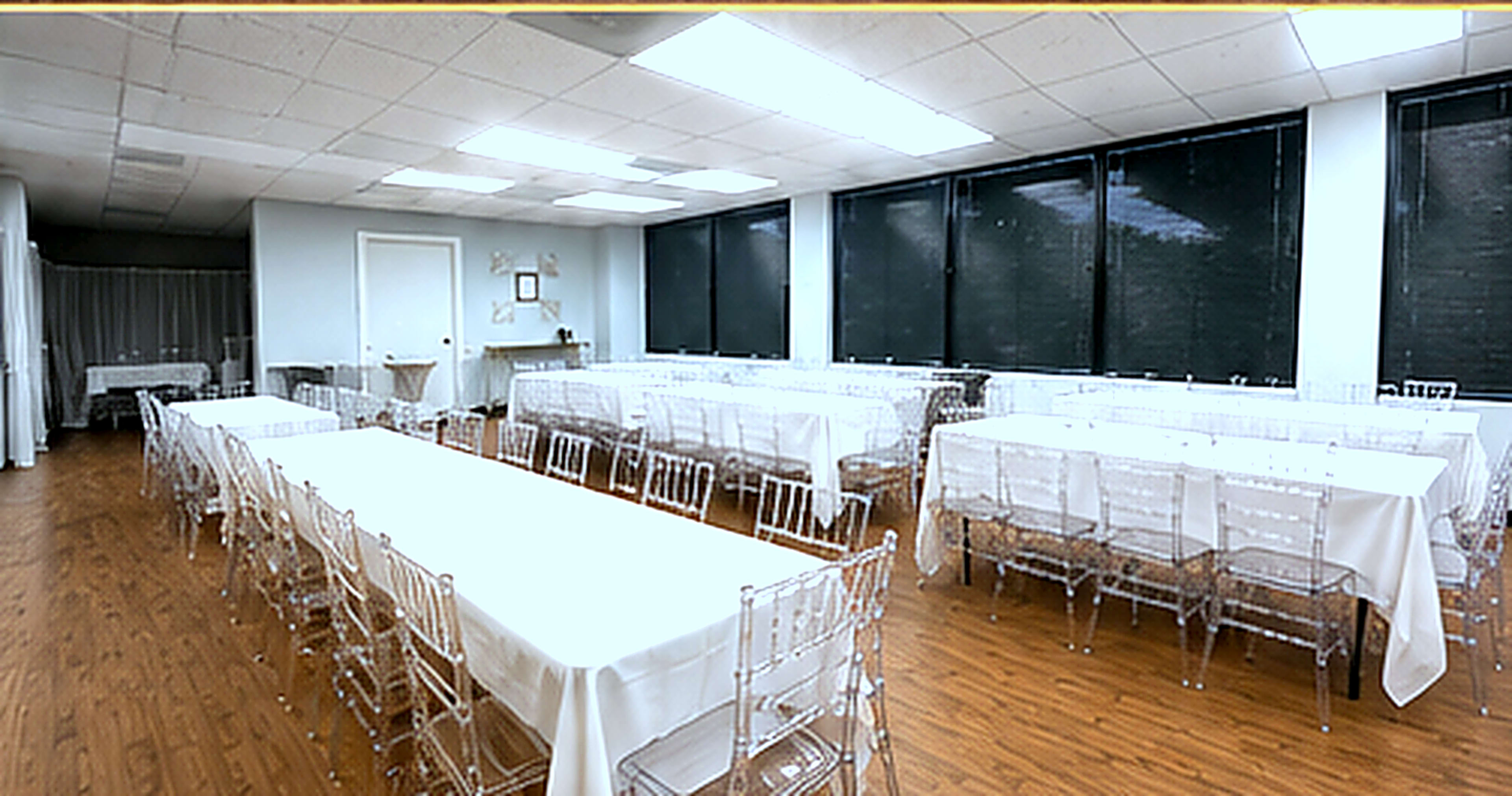 The image shows a spacious room with several long tables covered in white tablecloths and clear acrylic chairs arranged in rows.