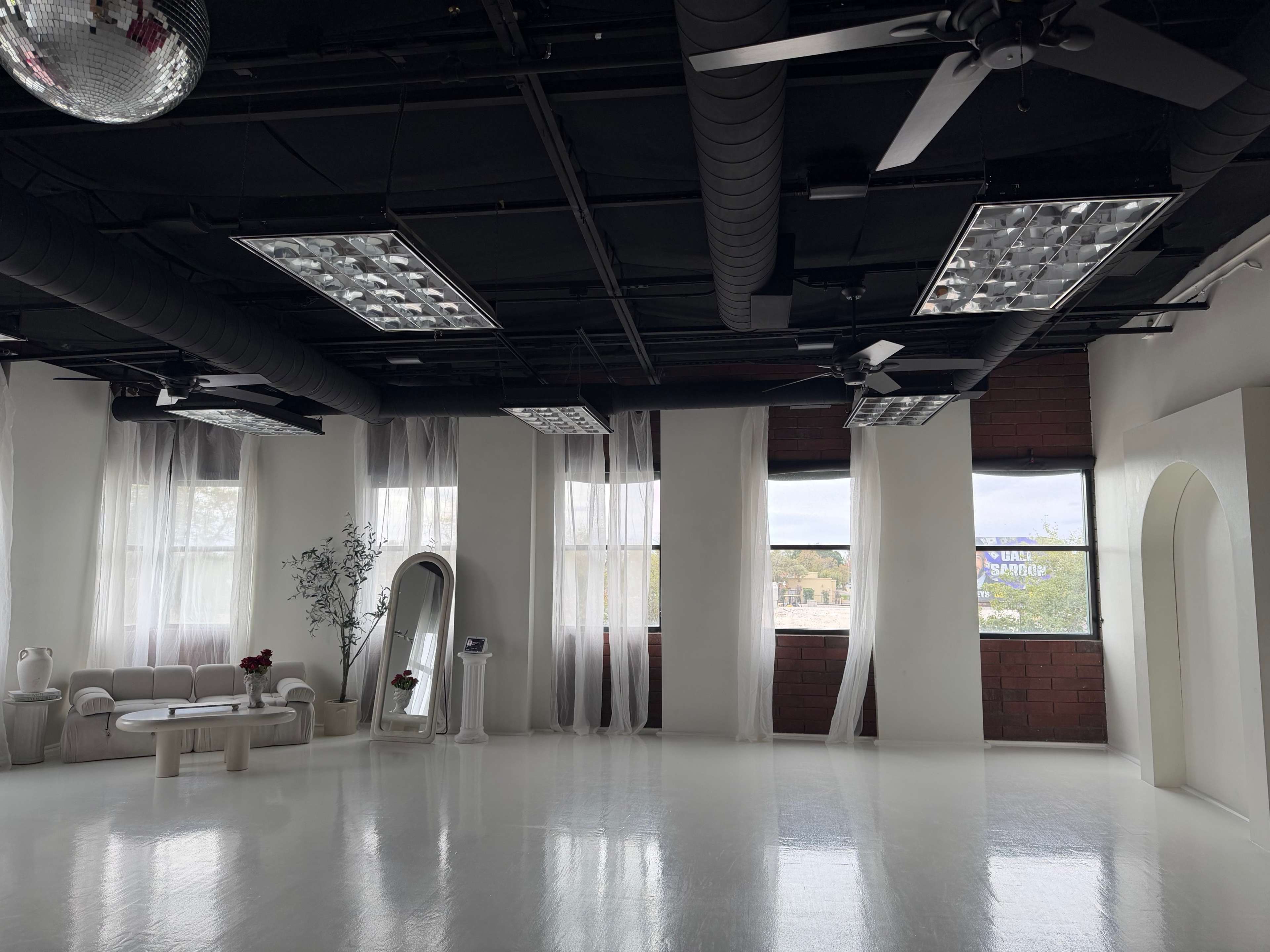 An empty, well-lit studio features white walls, a glossy floor, large windows, and minimalist furnishings.