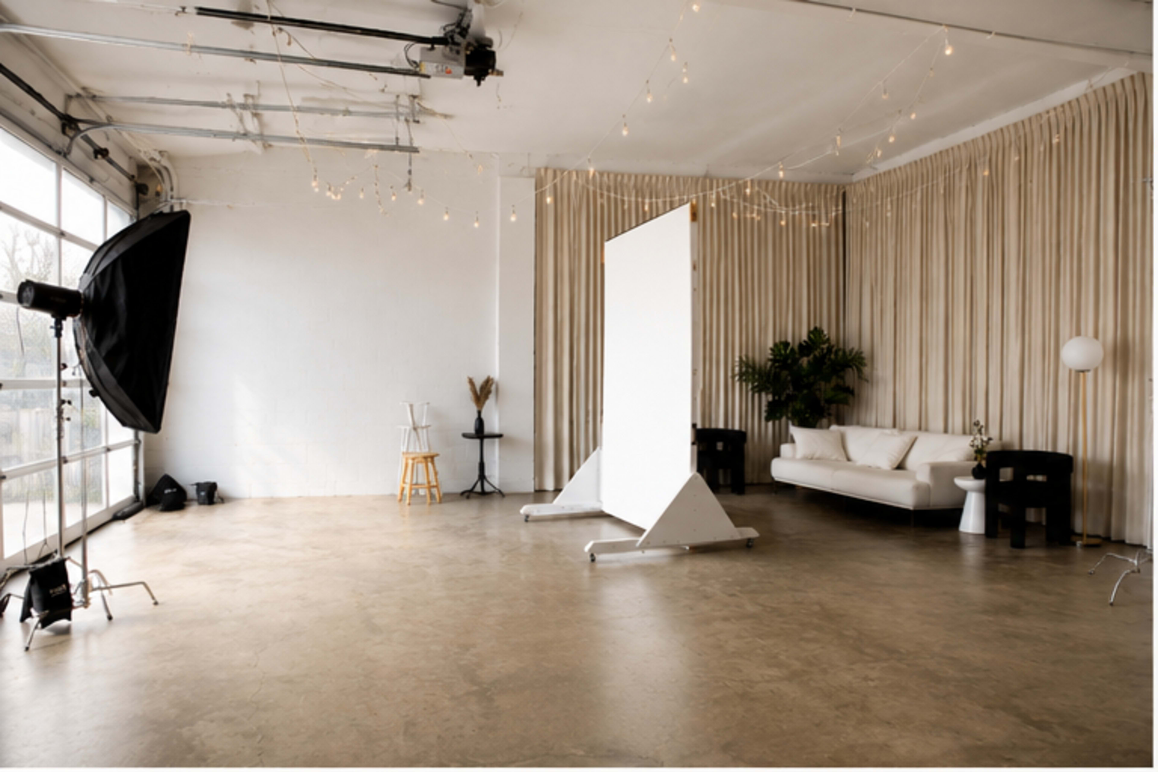 The image shows a spacious studio with a concrete floor, a large backdrop, soft lighting, a white sofa, and decorative elements like plants and string lights.