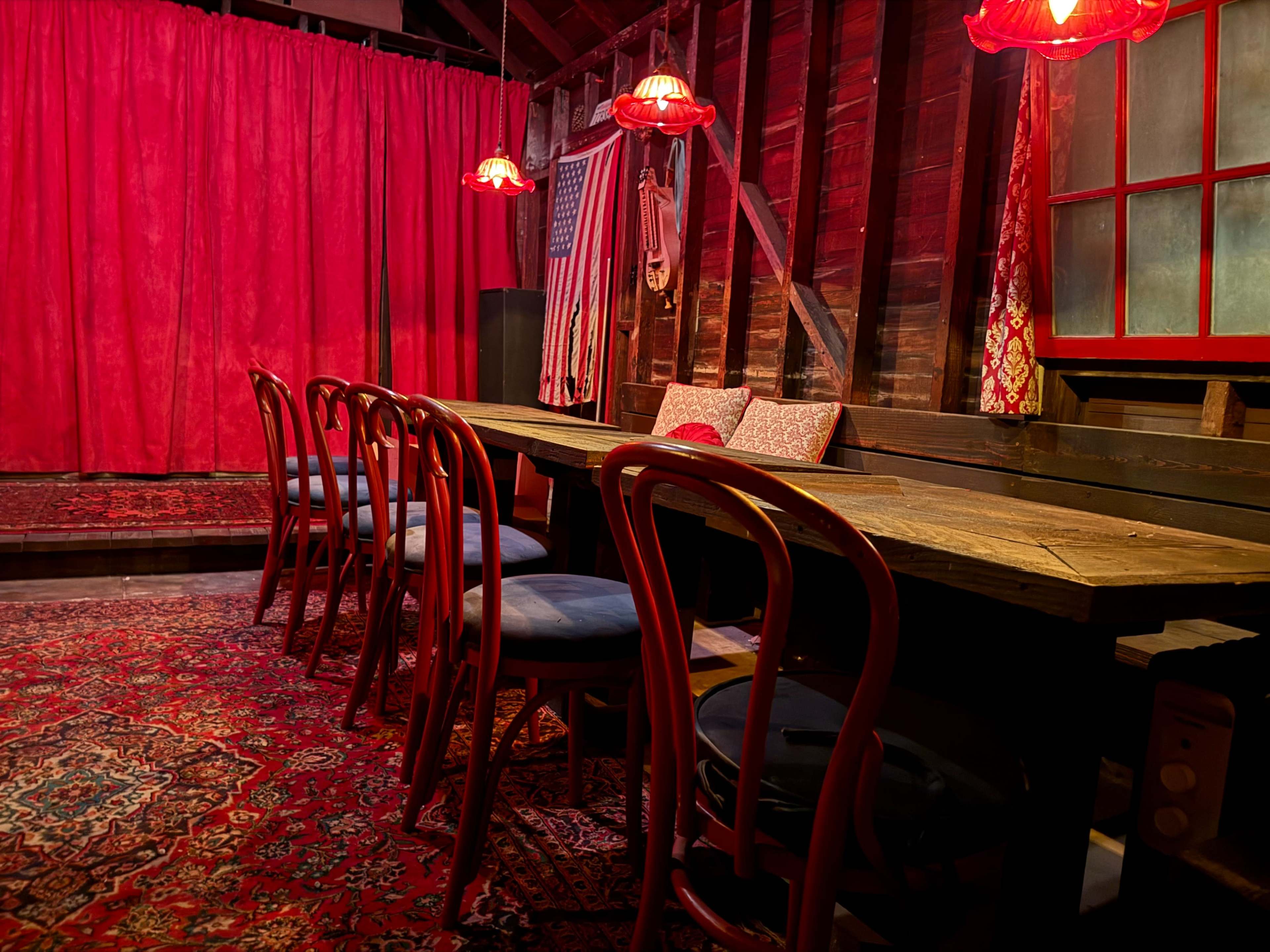 A long wooden table surrounded by chairs is set against a backdrop of red curtains and illuminated by red pendant lights in a rustic interior.