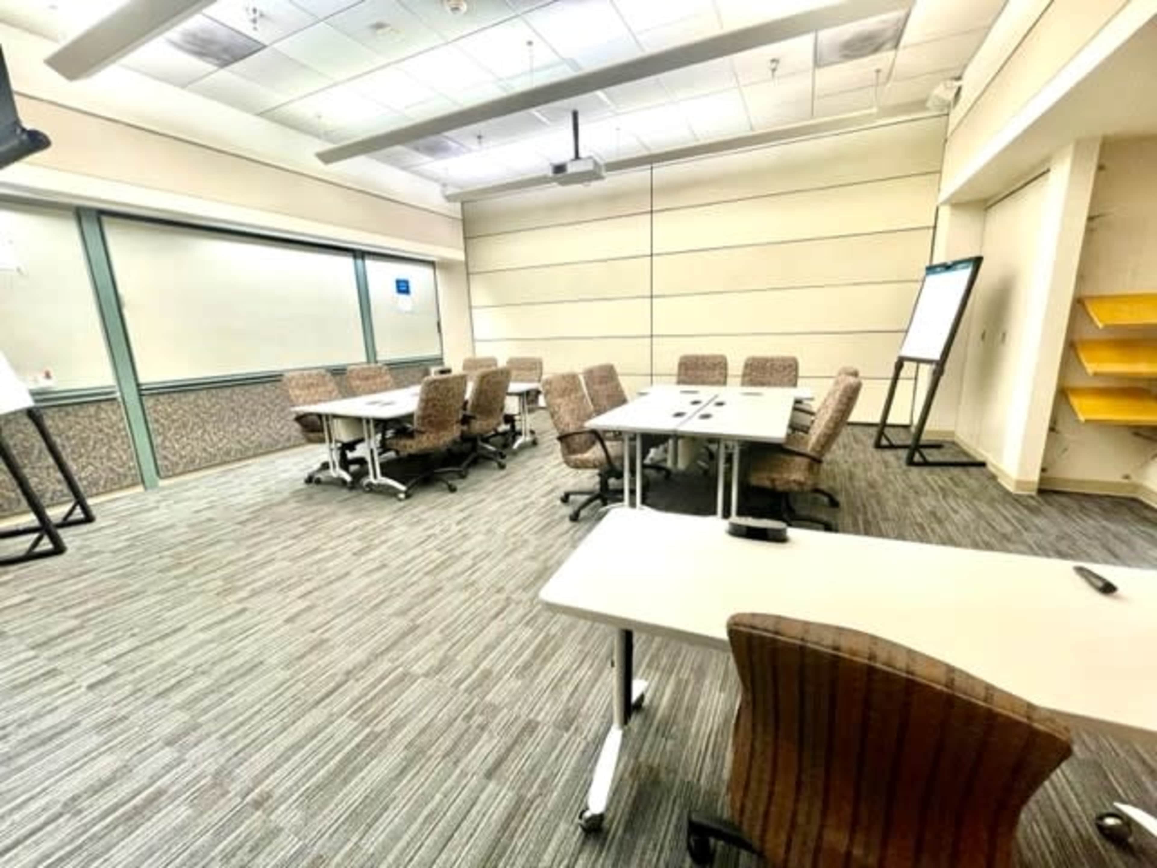 A conference room is organized with several tables and chairs arranged neatly around the space, featuring a projector and a whiteboard.