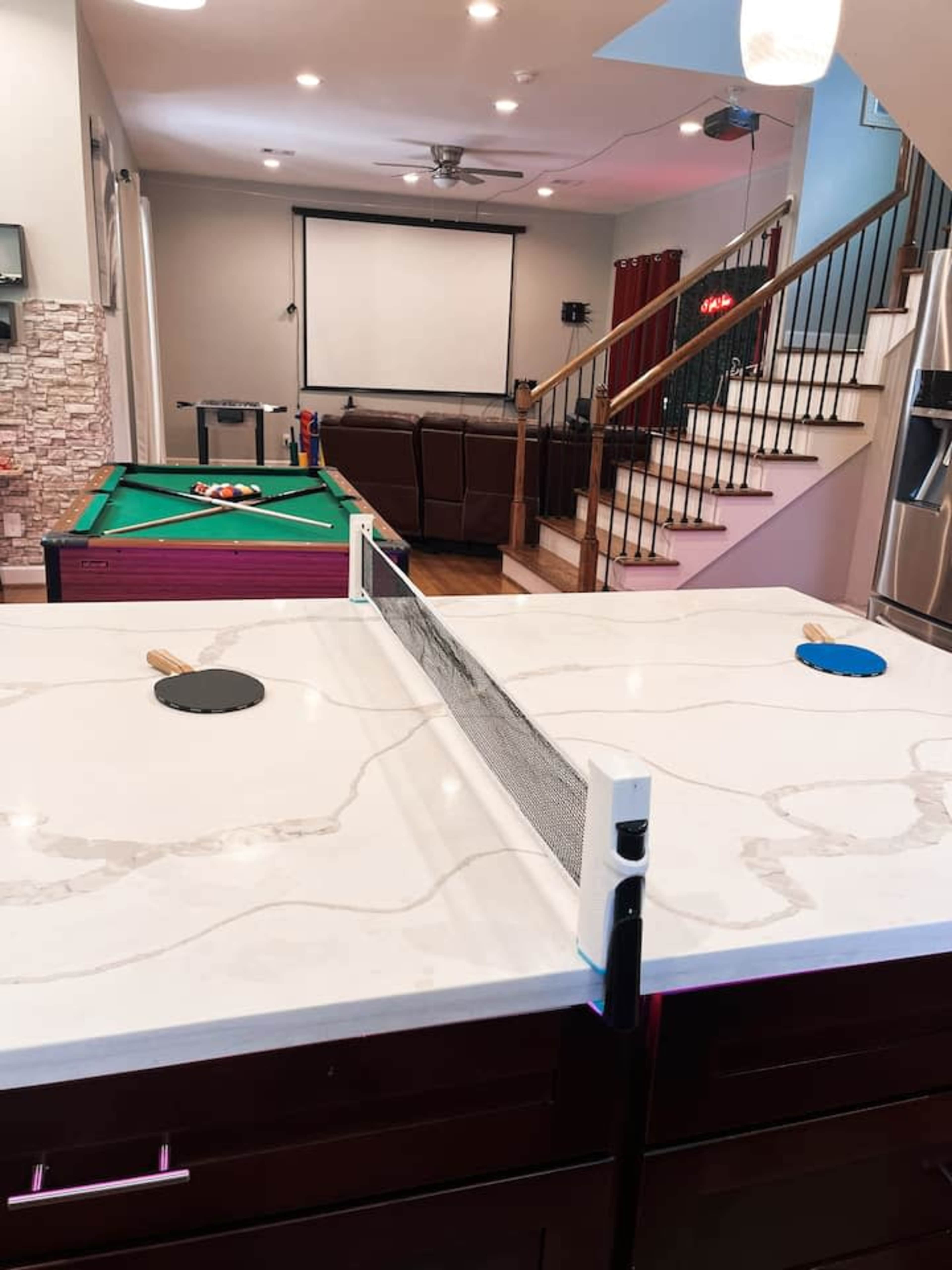 The image shows a recreational room featuring a ping pong table, a pool table, and a seating area visible in the background.