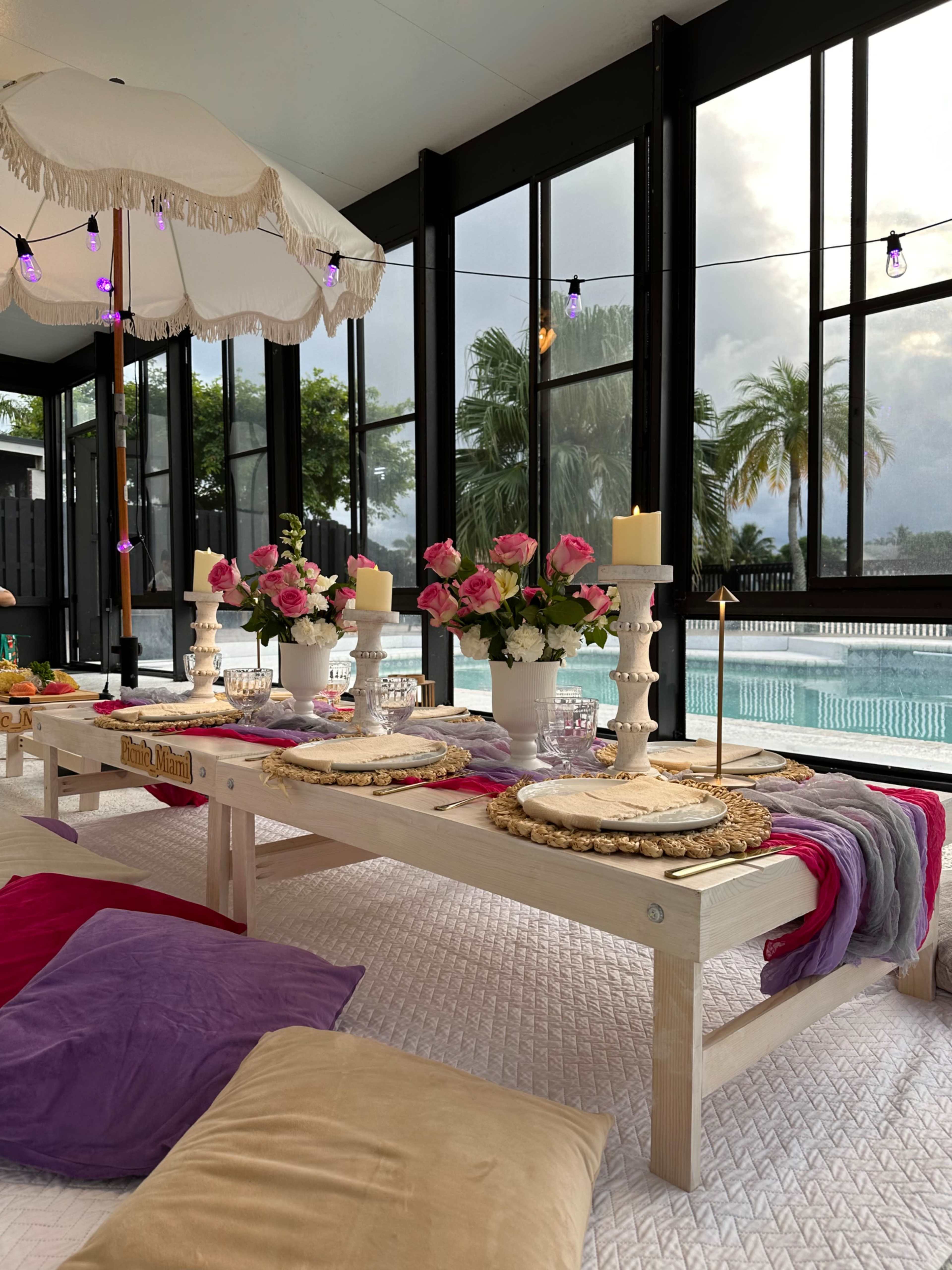 A beautifully arranged dining table with colorful cushions overlooks a swimming pool, surrounded by large windows and decor including flowers and candles.