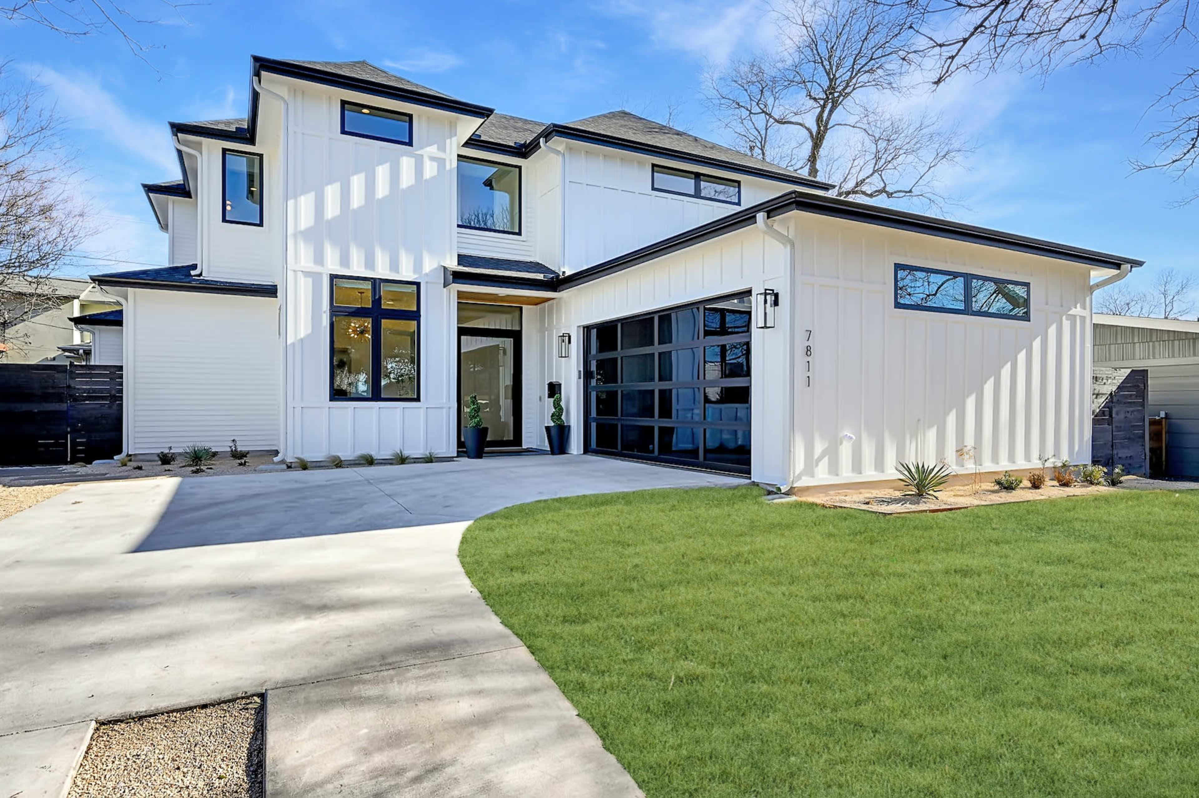 A modern white house with large windows and a manicured lawn, featuring a concrete driveway and minimal landscaping.