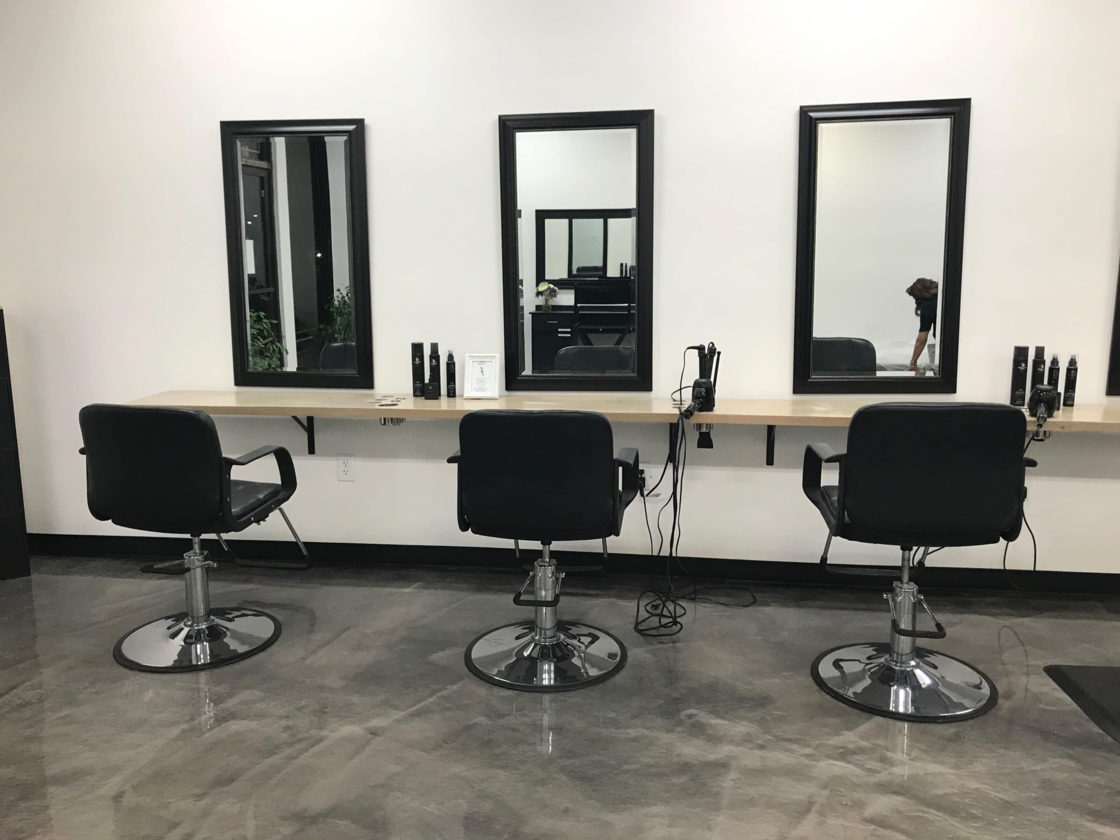 The image shows a modern hair salon interior with three black styling chairs positioned in front of mirrors on a wooden countertop.