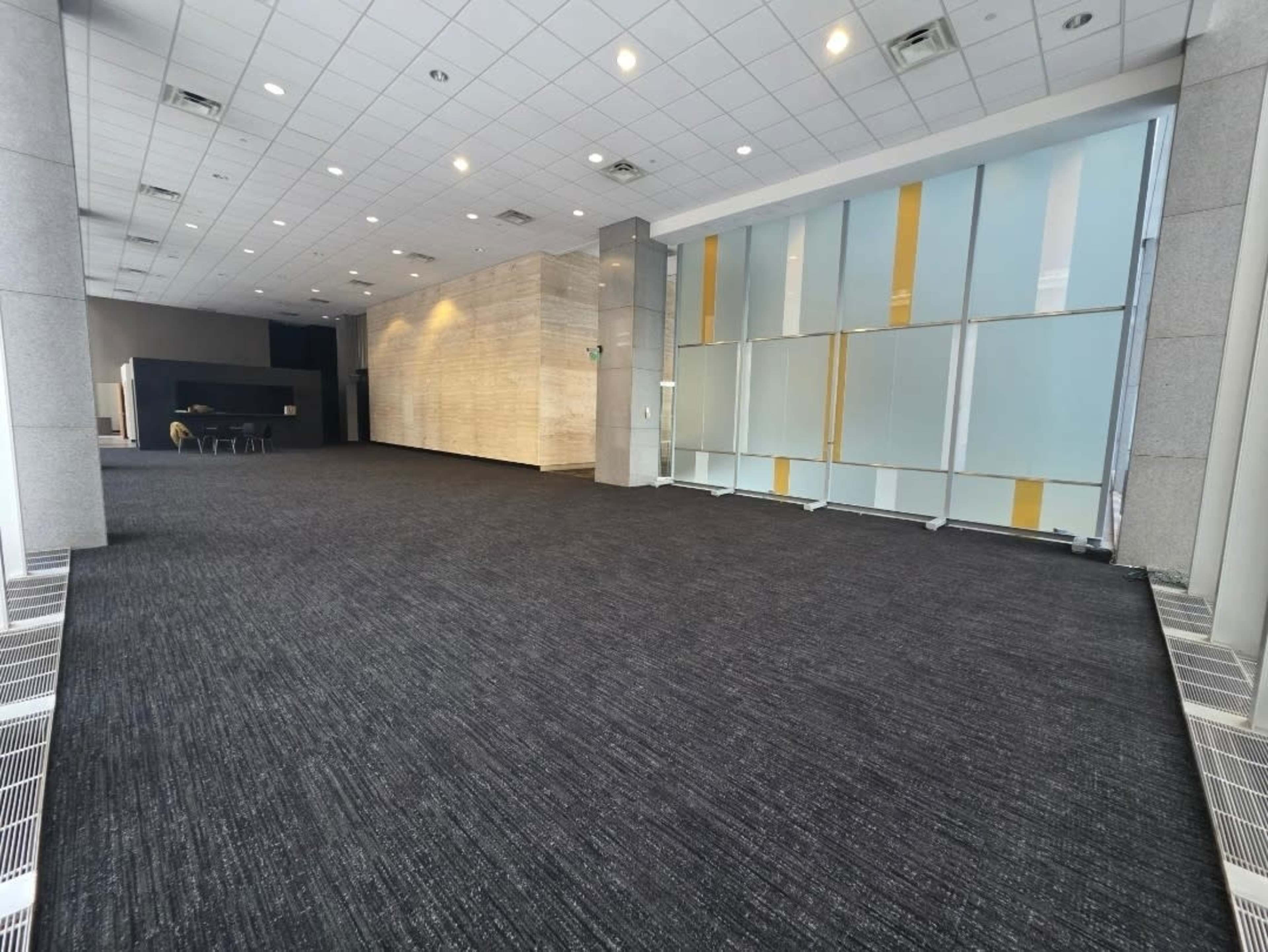 The image shows a spacious indoor area with gray carpet, featuring a partitioned section with large glass panels and soft lighting.