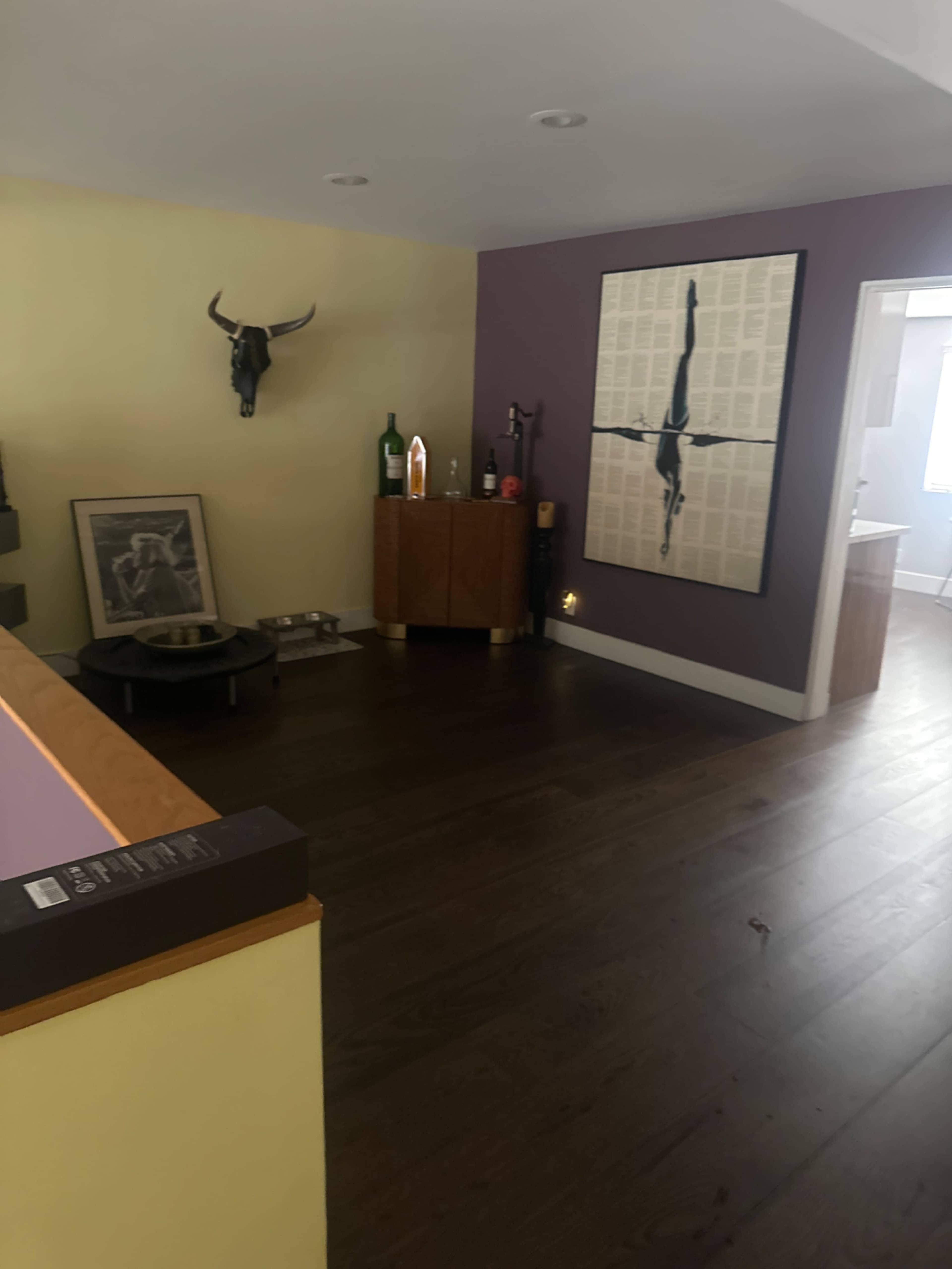 The image shows a room with wooden flooring, featuring a curved bar counter on the left, a bull head mounted on the wall, and a decorative cabinet against a purple wall.