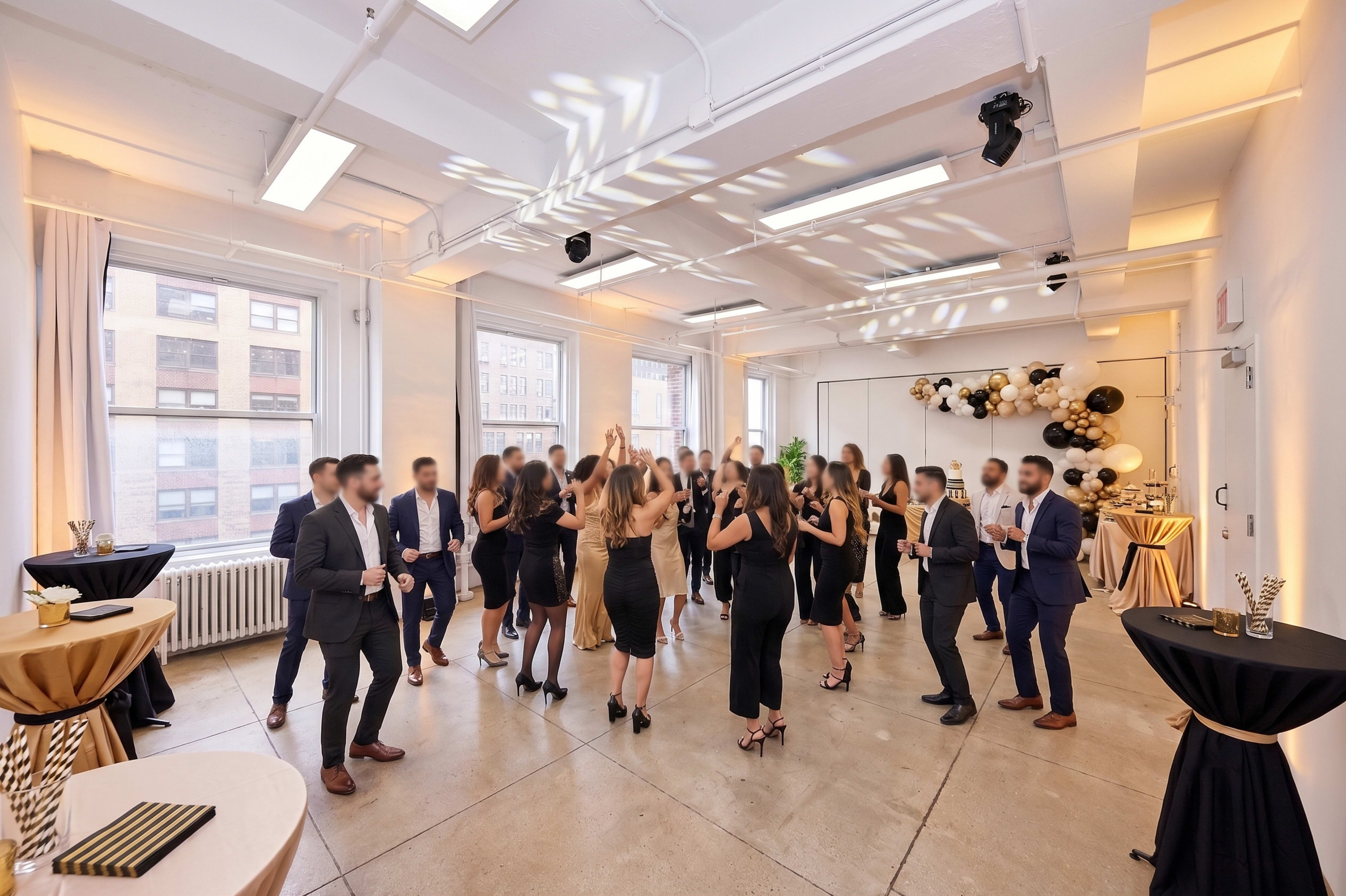 A group of people in formal attire are socializing and dancing at a spacious indoor event, decorated with balloons and ambient lighting.