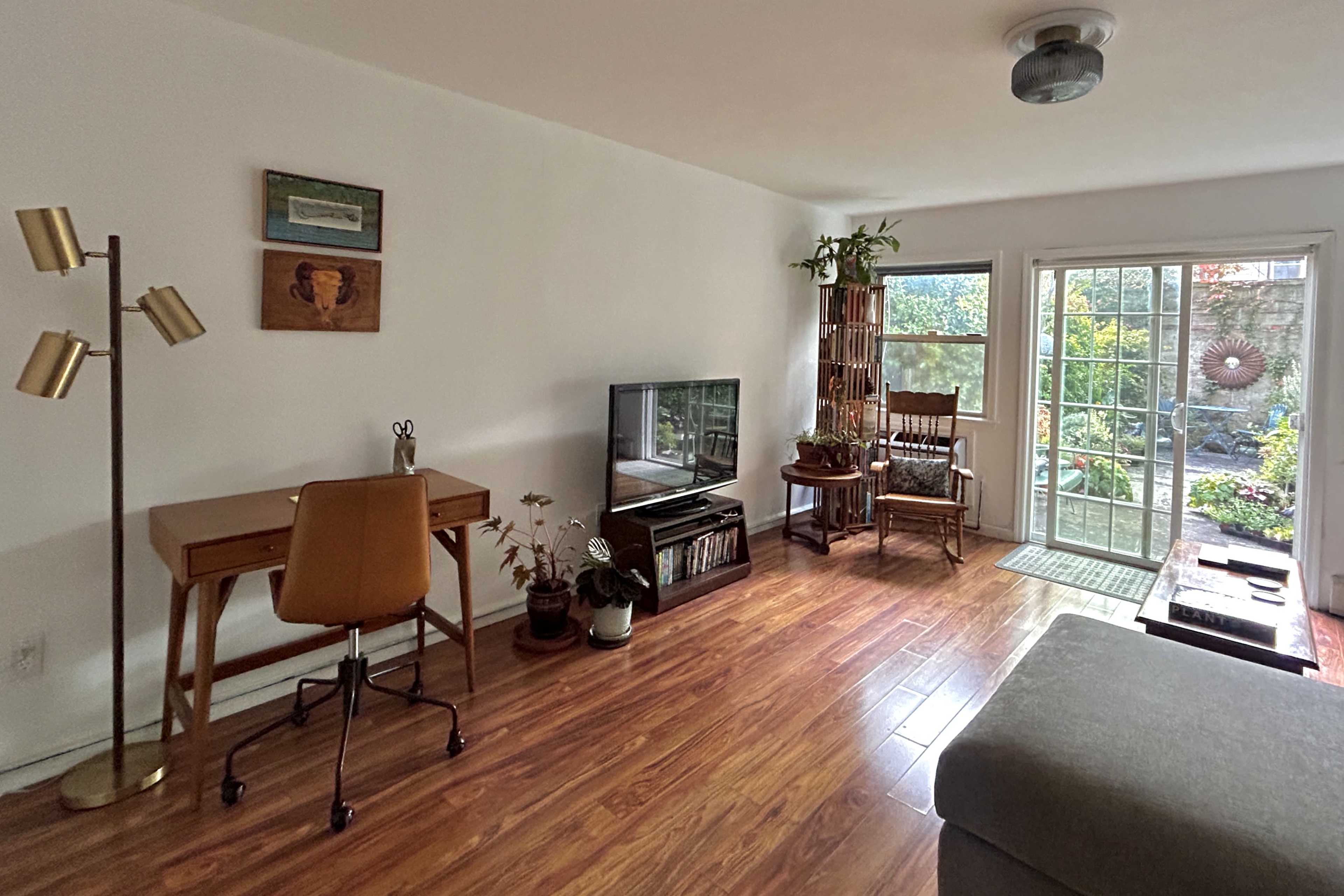 The image shows a bright living room with a desk, a television, and a seating area, all featuring wooden flooring and large windows leading to an outdoor space.