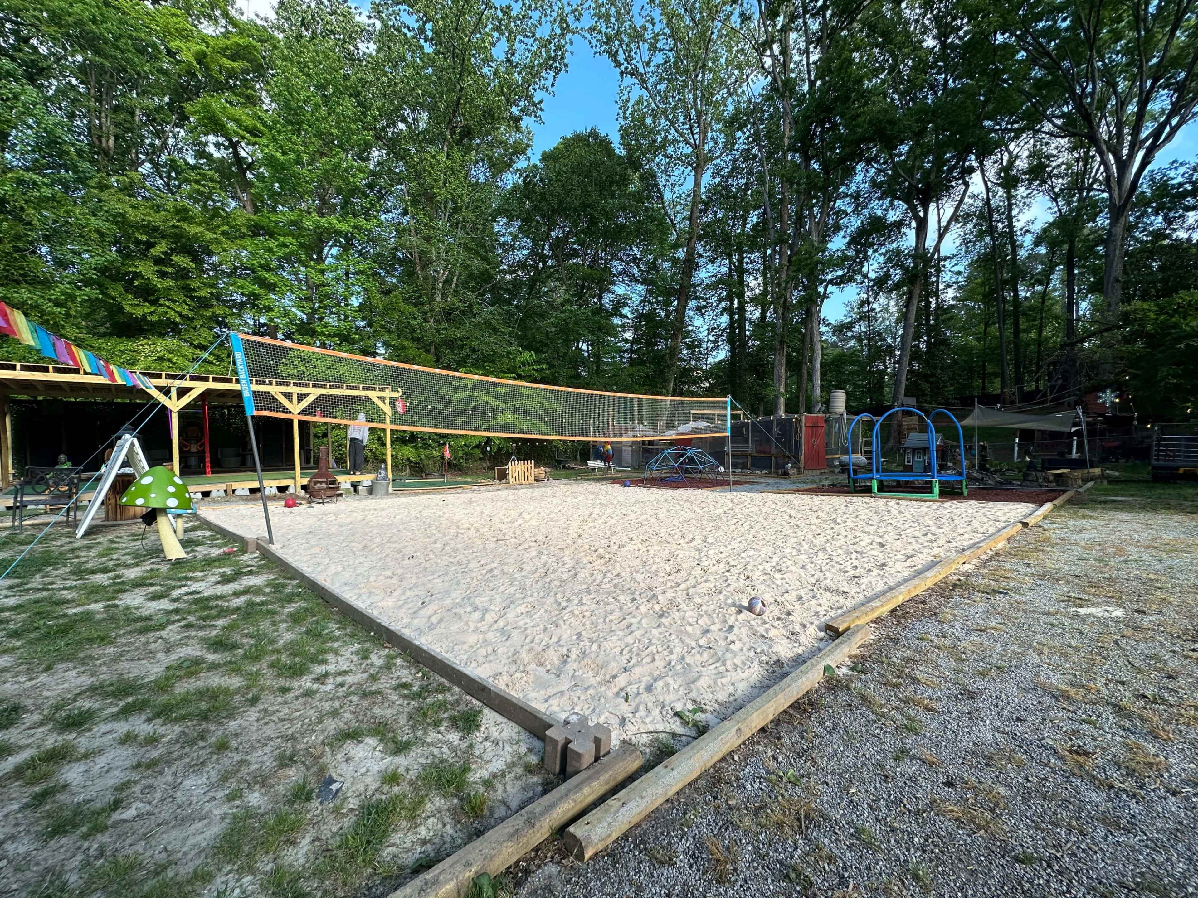 The image depicts a sandy volleyball court surrounded by trees, with playground equipment and benches nearby.